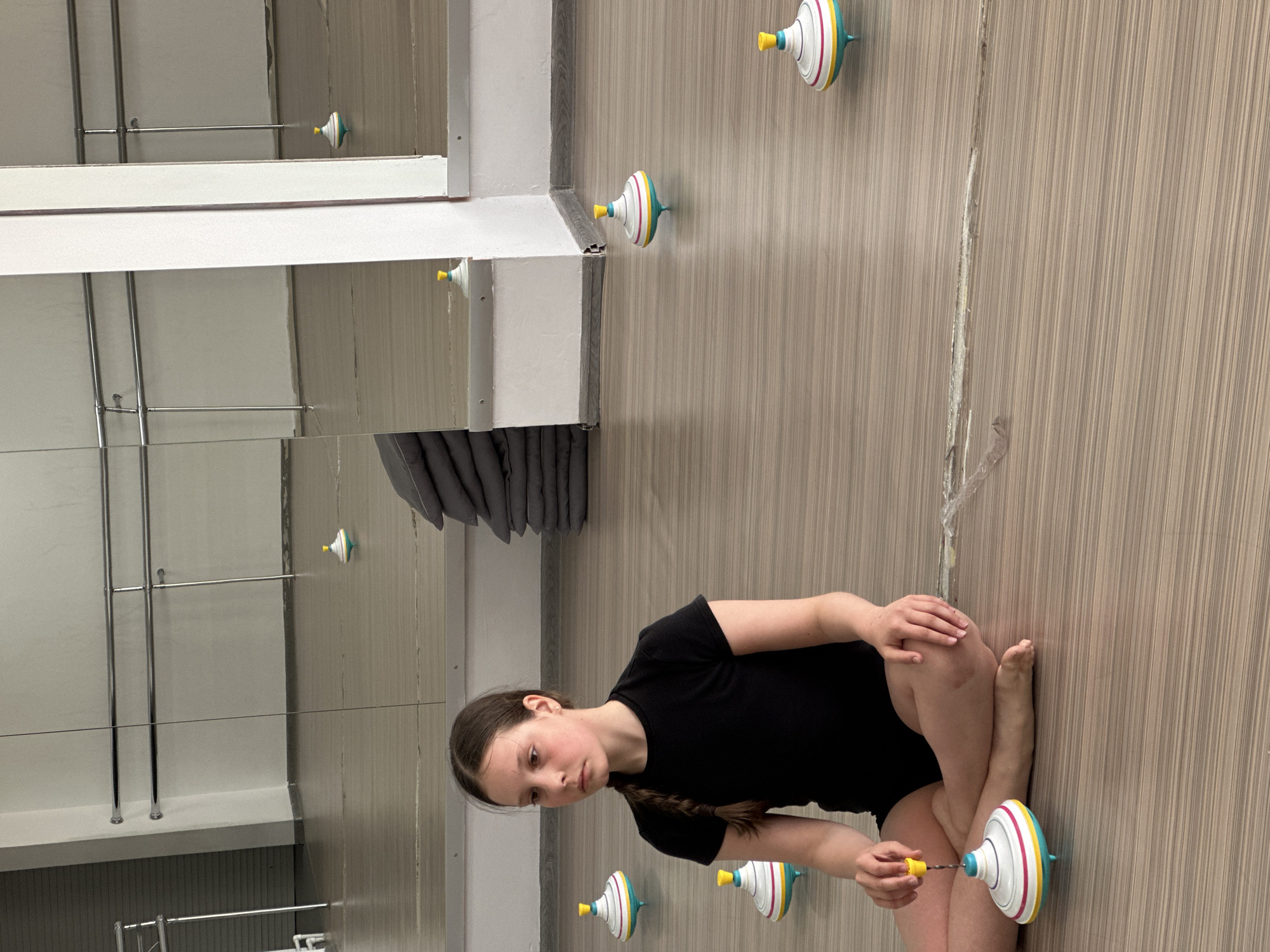 A sideways, eye-level indoor shot shows a young girl in a black shirt sitting cross-legged on a beige vertically striped floor. In her right hand, she holds a yellow and white peg over a spinning top of white, pink, yellow, and turquoise. One similar spinning top is behind her right foot, and two more are on the floor behind and to the left of her head. Another three are attached to the upper wall, one directly behind her head and two more between a white-framed mirror in the upper left and the wall. The mirror reflects a clothes-hanging rack and the three spinning tops. A crack and residue streak vertically along the wall behind the girl and beside the mirror. The city of Votkinsk, Russia, is not discernible from the information in the image.