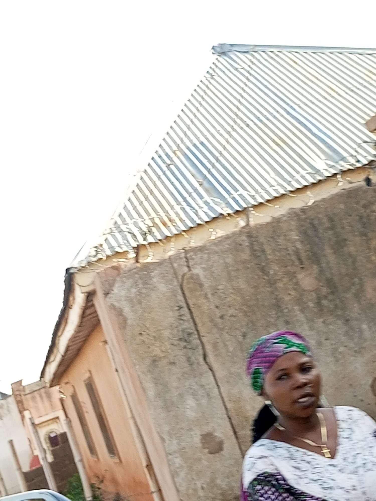 In the city of Gombe, Nigeria, a woman is captured in a medium shot, standing in front of a building with a corrugated metal roof. She is wearing a headscarf with purple and green patterns, gold hoop earrings, and a gold necklace with a pendant. Her attire includes a white top with what appears to be a patterned hem. The background shows a textured, light-colored wall and an adjacent building with windows. The sky above is a bright, overcast white.