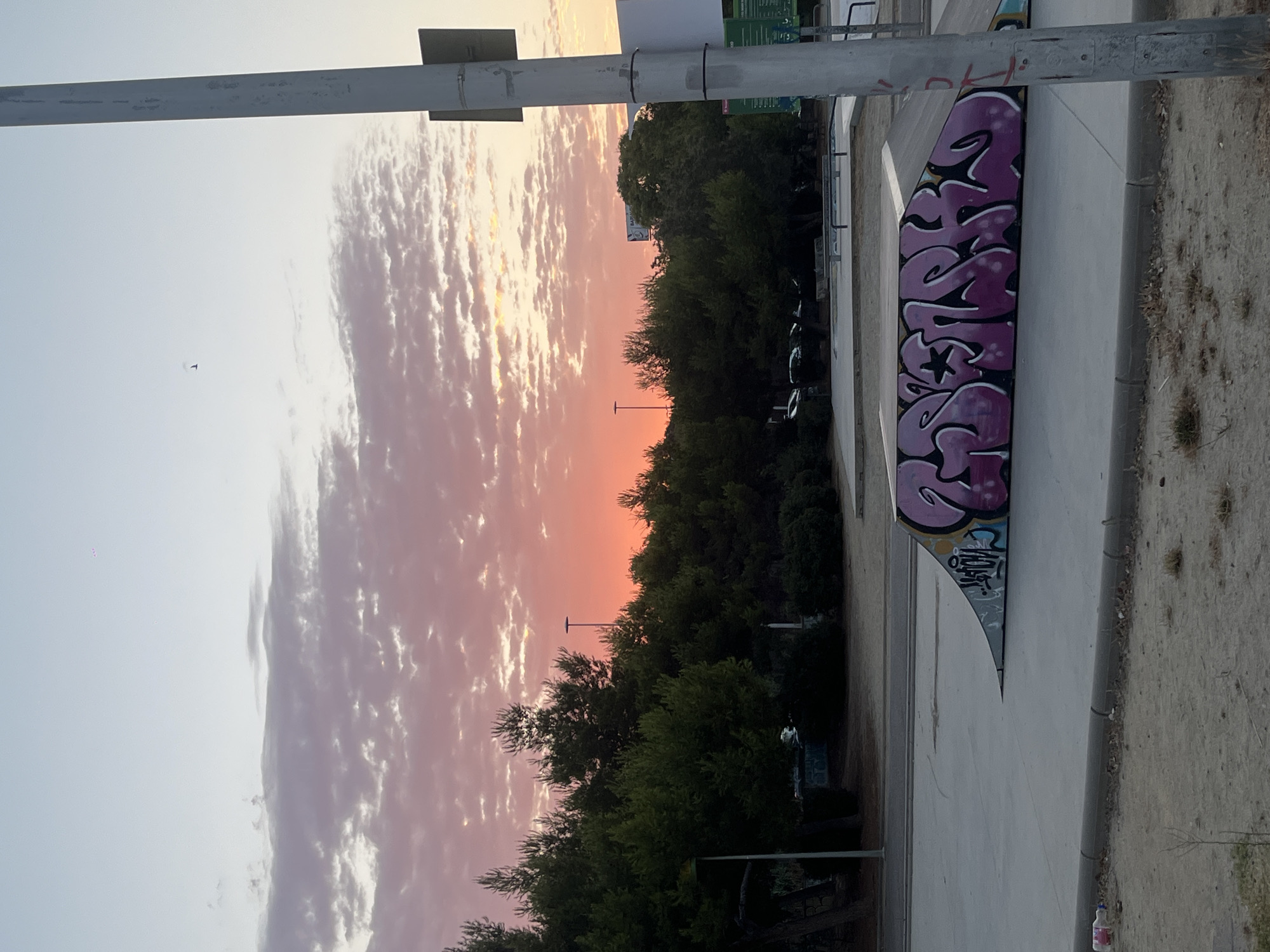 The image shows a sunset over a skatepark in Tarragona, Spain. The sky is filled with colorful clouds in shades of pink, orange, and yellow. In the foreground, there is a skate ramp with graffiti on it. The graffiti is in purple and black and appears to be a stylized word or name. There are trees and streetlights in the background. A utility pole is on the right side of the frame.