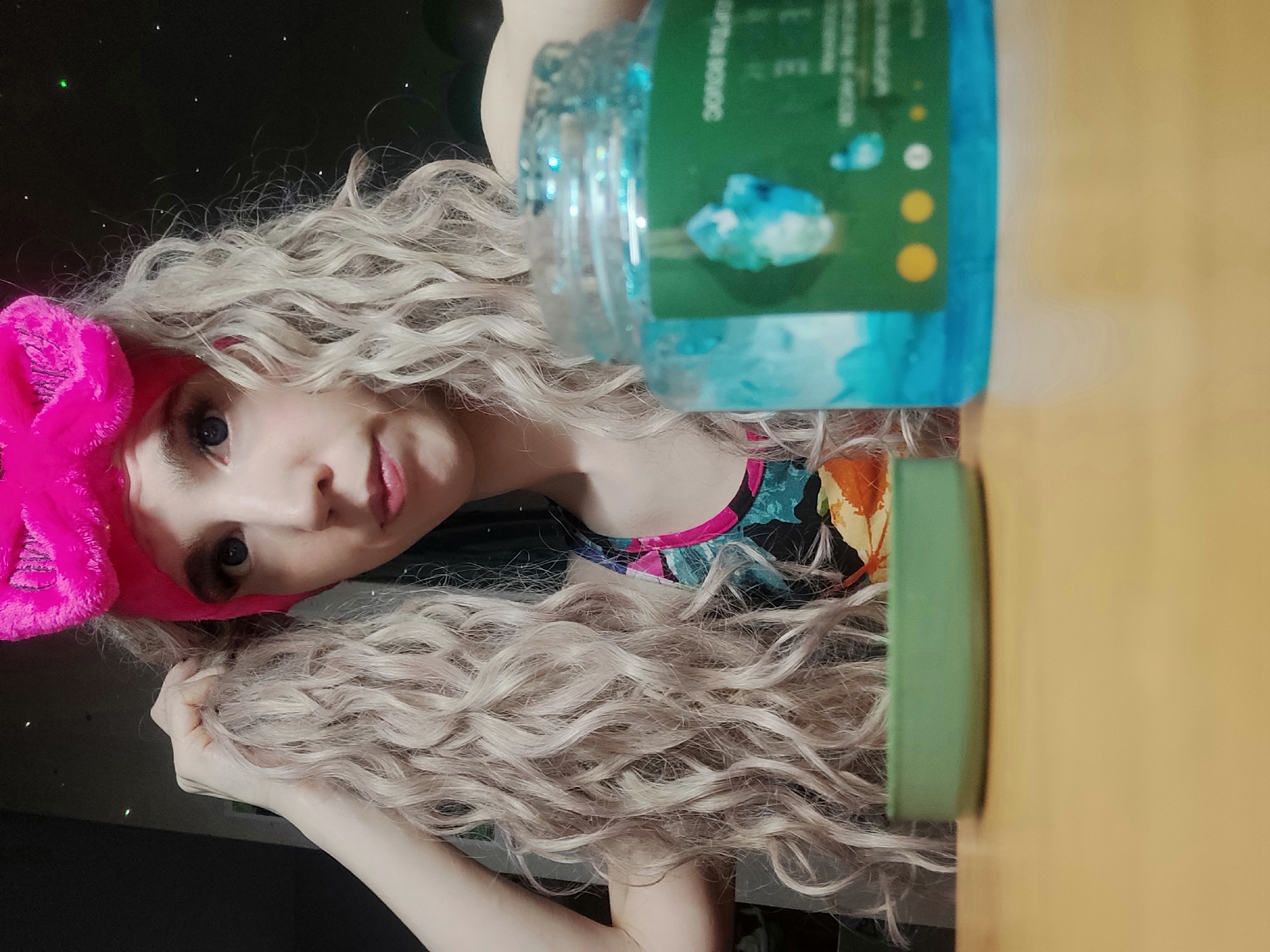 A woman with light skin, long curly blonde hair, and a pink headband is lying on a surface. She is wearing a multi-colored top. A clear plastic jar filled with a blue liquid and a green container are positioned near her. The background is dark. The location is Chaikovsky, Russia.
