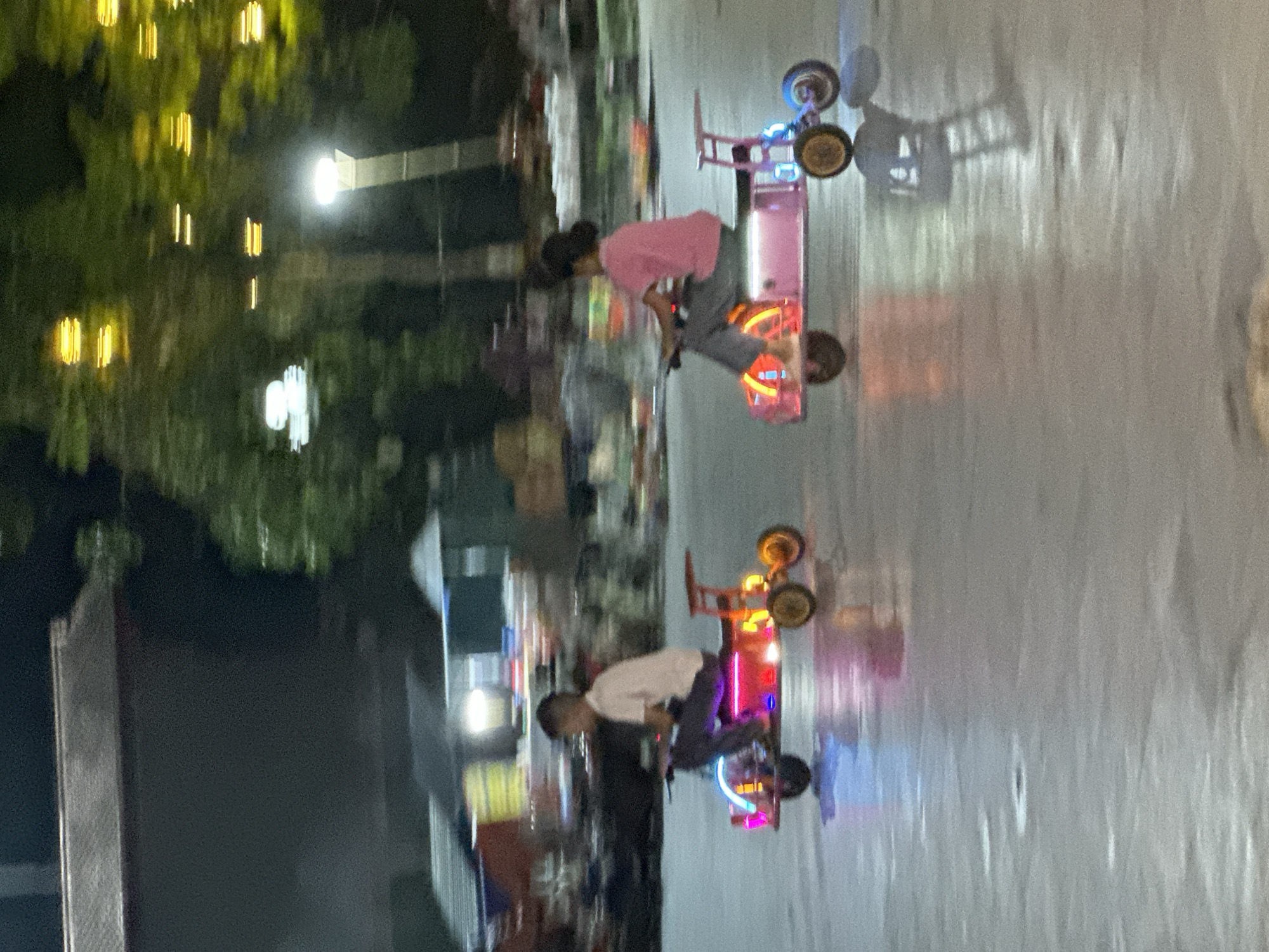 The image depicts an outdoor night scene featuring two individuals, likely children or young adults, riding brightly illuminated recreational vehicles on a reflective surface.
**Subjects:** Two people are central to the image. One individual, wearing a pink shirt and grey pants, is riding a pink vehicle. Another individual, dressed in a white shirt and dark pants, is operating a red and orange vehicle. Both appear to be focused on their activity.
**Objects:** The main objects are the two small, multi-wheeled recreational vehicles, which resemble go-karts or pedal cars. These vehicles are extensively decorated with vibrant, multi-colored lights (pink, blue, red, orange), illuminating the surrounding area and creating noticeable reflections. In the background, there are trees or dense foliage, adorned with yellow or orange string lights. A street lamp or similar white light source is also visible.
**Scene and Setting:** The scene is set outdoors at night. The ground surface is highly reflective, appearing wet, possibly from recent rain, or polished, creating clear, elongated reflections of the vehicles, their lights, and the overhead lighting. The background is blurred but suggests an urban or semi-urban environment with dark silhouettes of buildings and illuminated structures.
**Activity or Event:** The individuals are engaged in riding these light-up vehicles, suggesting a recreational or leisure activity, possibly in a park, plaza, or designated play area.
**Notable Details:** The time of day is clearly night, emphasized by the darkness and the reliance on artificial light sources. The wet or highly reflective ground is a prominent feature, enhancing the visual appeal with dynamic reflections. The vehicles' colorful lights create a lively and festive atmosphere. There is no visible text in the image.