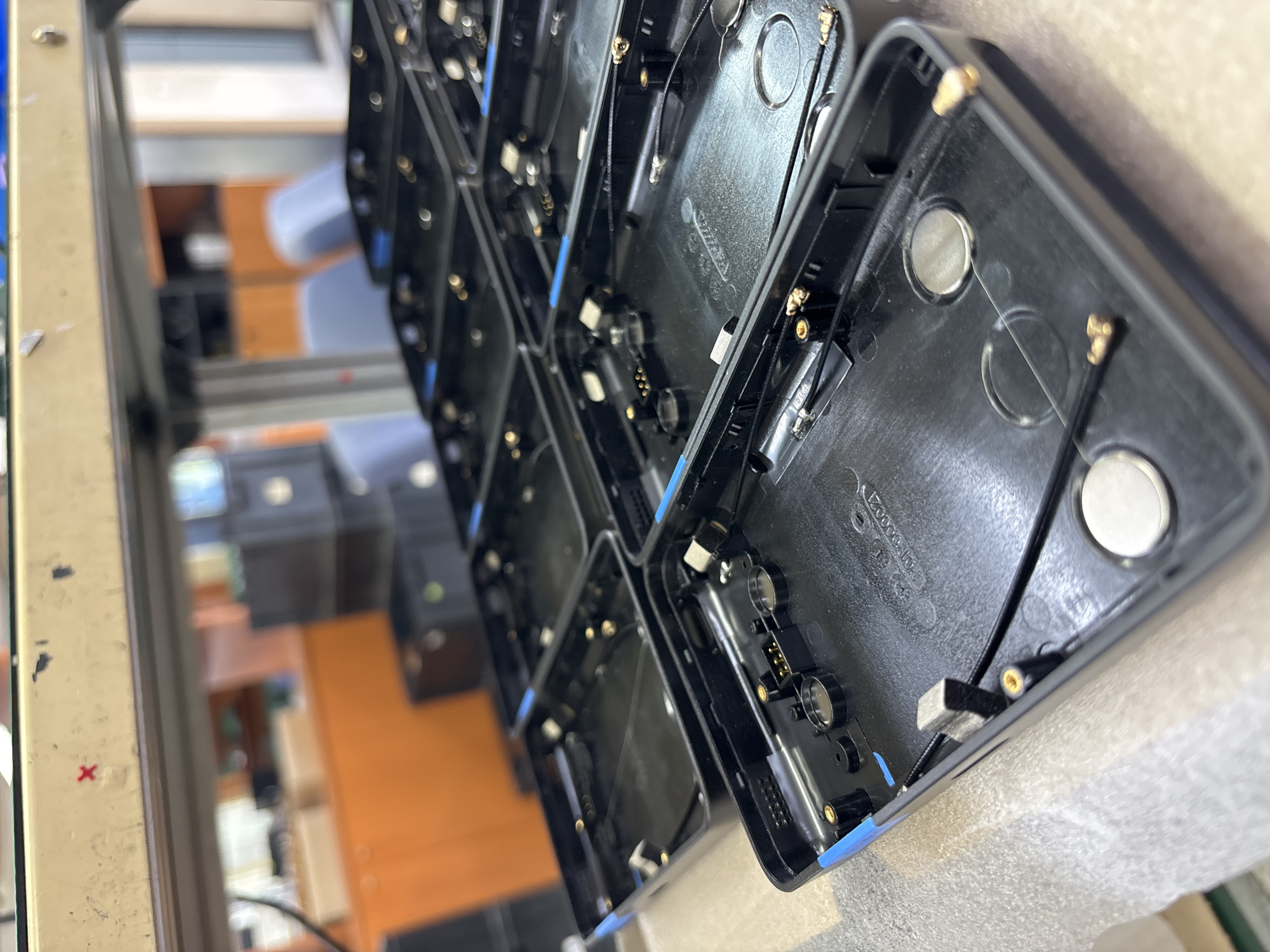 The image shows a row of black electronic device housings, possibly smartphone or tablet cases, arranged on a workbench. The interiors of the housings are visible, revealing various electronic components and connectors. The housings are of similar design. The background is blurred and appears to show a workshop or assembly area with equipment and furniture. It is impossible to determine the specific location in Sofia, Bulgaria, or the presence of people or any specific actions without additional context.