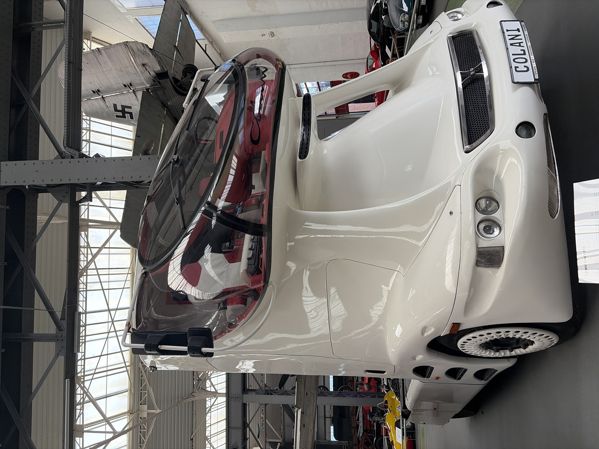 In the city of Speyer, Germany, inside a museum, a white, futuristic truck named "COLANI TRUCK MB" is on display. The truck has a unique, bulbous white cab with a transparent bubble-like roof, revealing a red and white interior with seats. The front of the truck features a prominent grille, rounded headlights, and a license plate that reads "COLANI". In the background, other vehicles, including a yellow race car and a dark green car, are visible. Above the truck, an airplane with a swastika symbol on its tail is suspended from the ceiling. The museum's architecture includes a high, industrial-style ceiling with exposed beams and large windows.