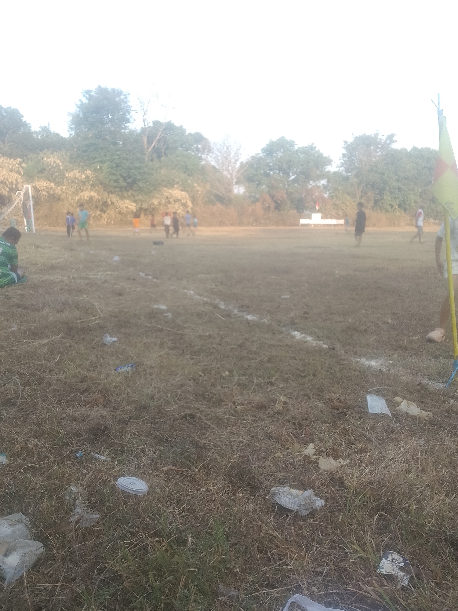 The image depicts an outdoor scene on a dry, grassy field, likely a sports ground, with a soccer goal visible on the left. Several individuals, appearing to be young people, are scattered across the field, some in motion, suggesting a game or practice is in progress. The ground is littered with various pieces of trash, including plastic bags and a white lid. In the background, a line of trees forms a natural boundary. A yellow and red flag on a pole stands prominently in the right foreground, potentially marking a corner of the field. The overall lighting suggests it is daytime, possibly late afternoon or early morning due to the warm hue of the light and long shadows. There is no specific visual information in the image that definitively places it in Kecamatan Haurgeulis, Indonesia; the scene is generic to many outdoor sports fields.