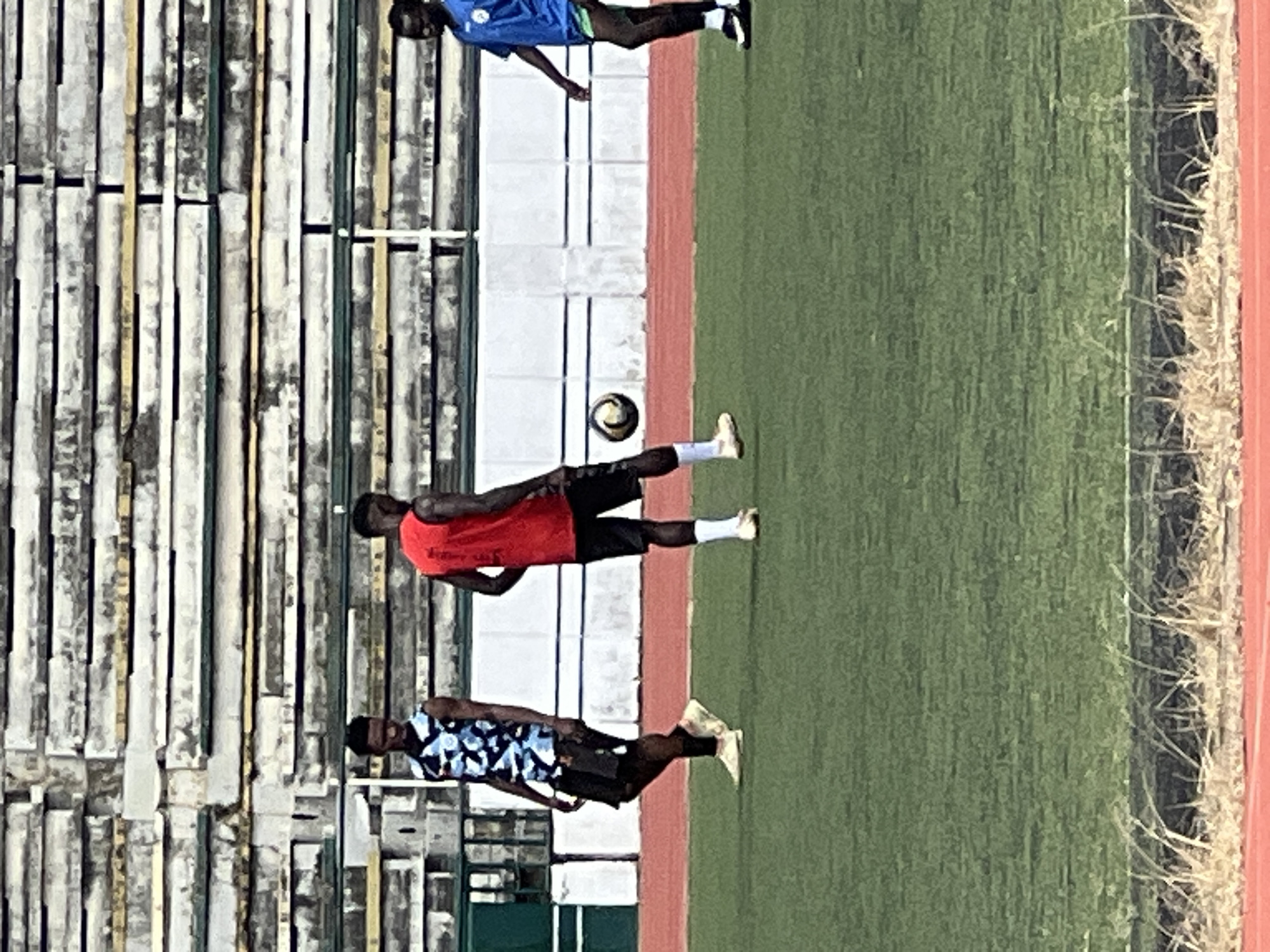 A group of three young Black men are on a green astroturf field, likely a sports stadium, in Rafin Par, Nigeria. The man on the left wears a sleeveless blue and black patterned jersey and black shorts. The man in the center wears a red sleeveless jersey and black shorts. The man on the right wears a blue jersey with a white emblem and green shorts. A soccer ball is visible between the legs of the center and right-most men. Behind them are weathered, tiered stadium seats with metal supports. The ground surface transitions from the astroturf to a red running track and then to dirt and dry vegetation along the edge of the field.