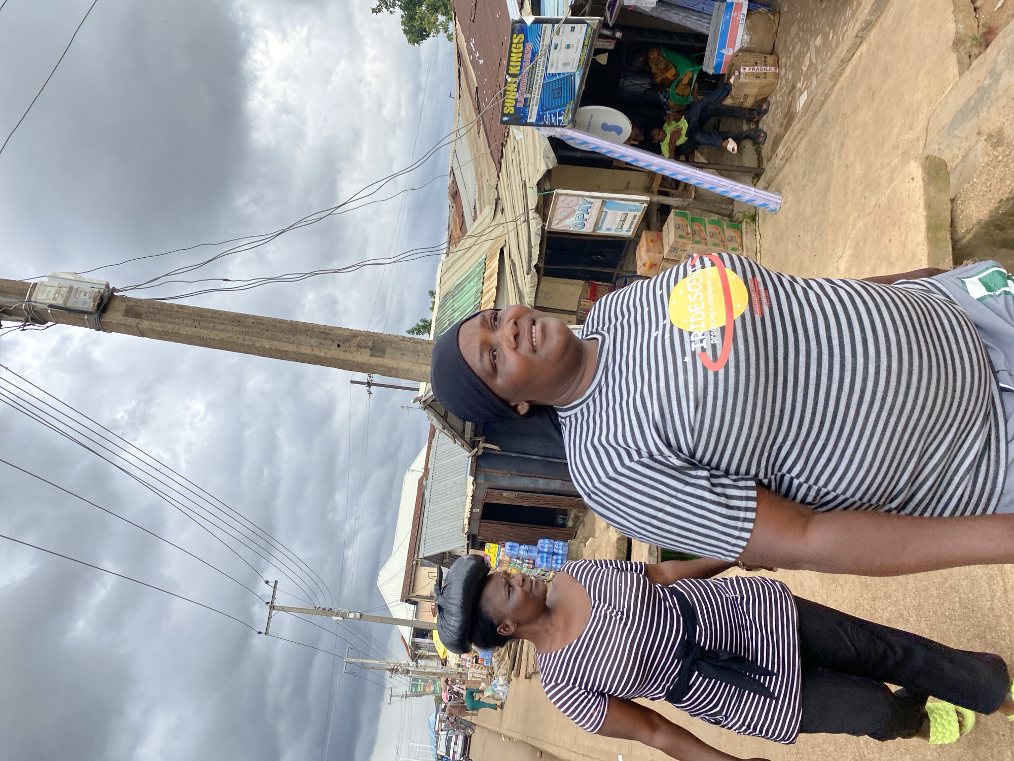 In Bukuru, Nigeria, two women are captured mid-stride on a street lined with makeshift stalls and overhead power lines. The woman in the foreground, wearing a black and white striped t-shirt and headscarf, has a yellow and red circular emblem on her shirt. To her left, another woman, also in a striped t-shirt, carries a large black plastic bag balanced on her head, while wearing a black sash around her waist and lime green slides on her feet.
The street is unpaved and appears dusty. Behind the women, a collection of modest structures with corrugated metal roofs house various goods, including bottled water and packaged items. A sign for "SUNNY KINGS ELECTRONICS HOLDINGS" is visible on one of the stalls. In the background, other individuals are present, some seated, and a car is parked on the side of the road. The sky is overcast with heavy clouds.