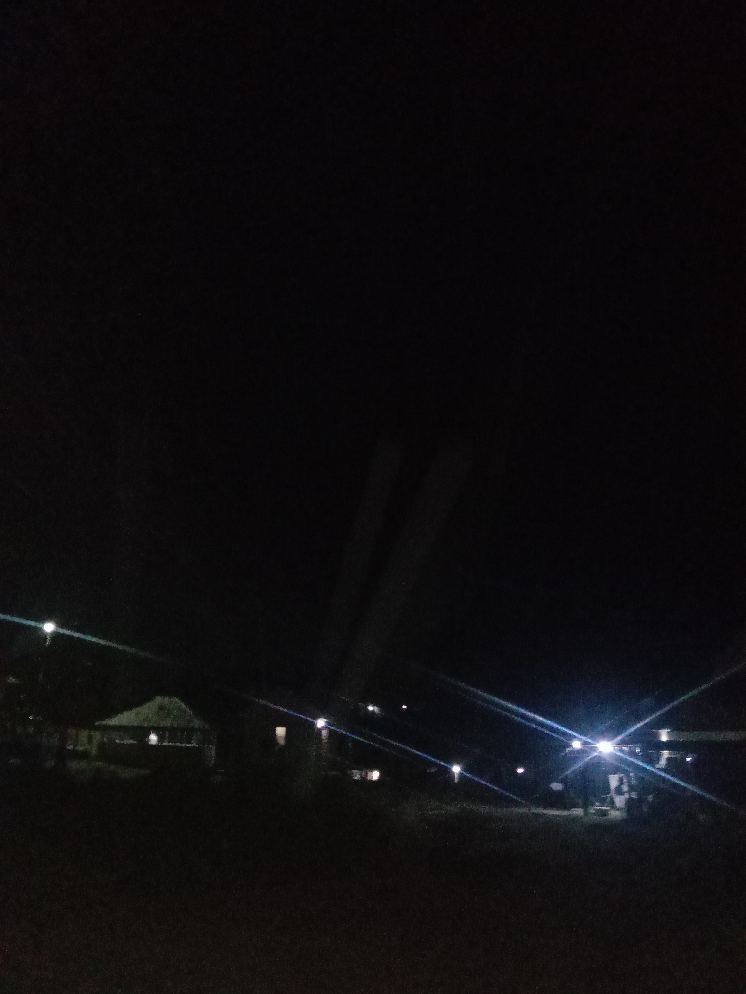 The image depicts a dark outdoor scene at night, illuminated by artificial lights. In the foreground, the ground is dark and appears to be unpaved. In the mid-ground, there are several structures. To the left, a building with a thatched roof is visible, with lights inside and around it. To the right, other structures are present, including what appears to be a gas station or a similar commercial establishment with bright lights and pumps. Several light poles with bright lights are scattered throughout the scene, casting beams and creating lens flares. The overall environment suggests a rural or semi-urban setting within the city of Lokoja, Nigeria, likely a roadside area with some commercial activity. There are no visible people in the image.
