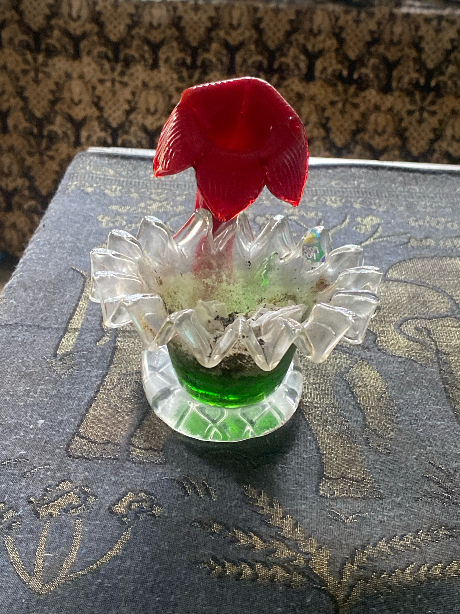 A close-up, high-angle shot shows a small, decorative glass item resembling a flower. The flower has a bright red, petal-shaped top and a clear glass base with ruffled edges and a green liquid or glass fill at the bottom. It sits on a dark, patterned fabric surface. The background is a brown and beige patterned textile. The image is taken indoors, with the lighting suggesting an artificial light source. There are no people or discernible actions in the image. This scene is not located in Batam, Indonesia.