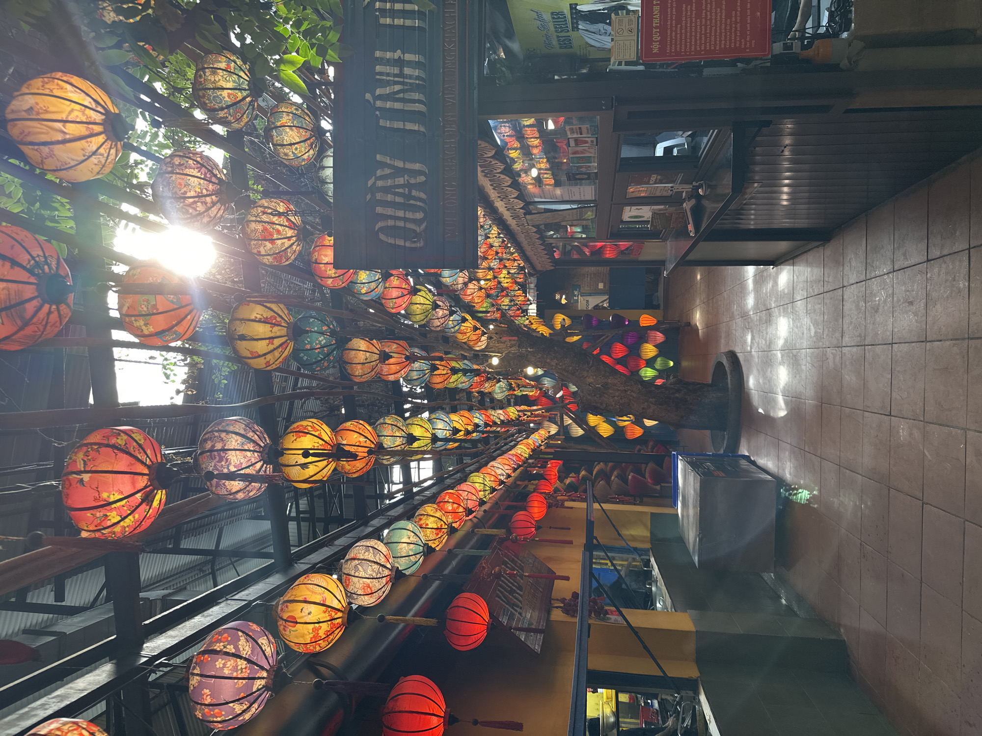 The image shows a narrow alleyway in what is likely a tourist district. Above, strung on wires or poles, are many traditional Vietnamese lanterns in various colors. A wooden sign is visible, and further down the alley, shops or businesses can be seen on both sides. The alley is tiled, and in the background, there are signs with Vietnamese text. Given the lanterns and text, the location could be in or near Đà Nẵng, Vietnam.