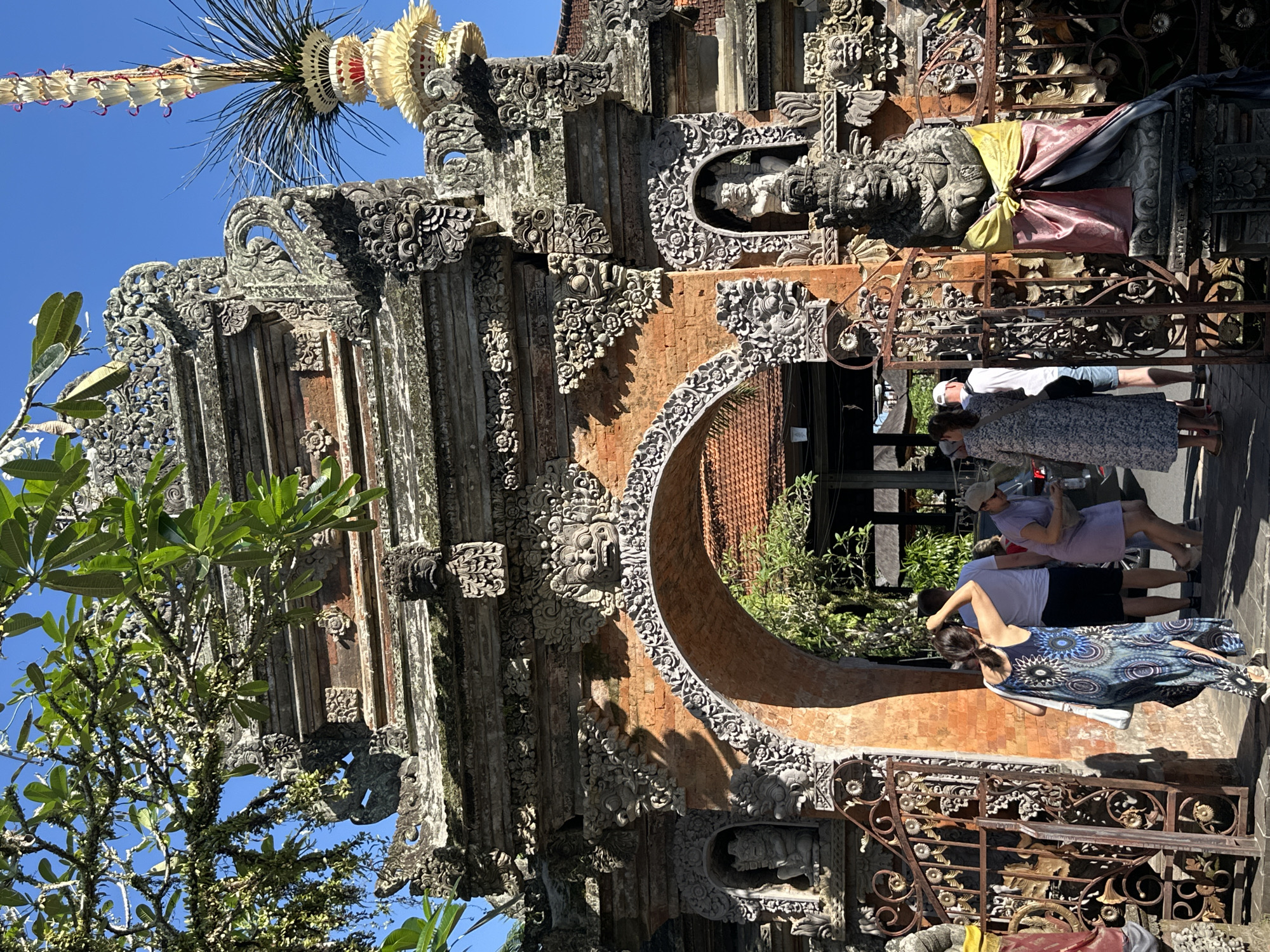 This image depicts an ornate stone gateway in Ubud, Indonesia, a district known for its rich cultural heritage. The gateway, constructed with intricately carved stone and brick, features multiple tiers of decorative elements, including facial motifs and floral patterns. An arched opening allows passage, and the entire structure is adorned with weathered textures indicative of age and exposure to the elements.
In the foreground, several individuals are visible. A woman in a dark blue and white patterned dress is walking away from the viewer, her back to the camera, with her right hand touching the back of her head. To her left, a man in a white t-shirt and blue shorts is partially visible. Further to the right, a woman in a light purple dress and a light-colored cap is interacting with someone off-camera, while another person in a light-colored dress is also present. Another statue, adorned with yellow and pink fabric, stands prominently to the right of the gateway's entrance.
The environment is bathed in sunlight, casting shadows and highlighting the details of the architecture. Lush green foliage, including tree branches with leaves, extends into the frame from the top left, partially obscuring the sky, which is a clear blue. Beyond the gateway, glimpses of a red-tiled roof and more greenery suggest a serene, possibly temple-like, setting. The overall scene conveys a sense of cultural immersion and architectural grandeur, characteristic of the Ubud region.