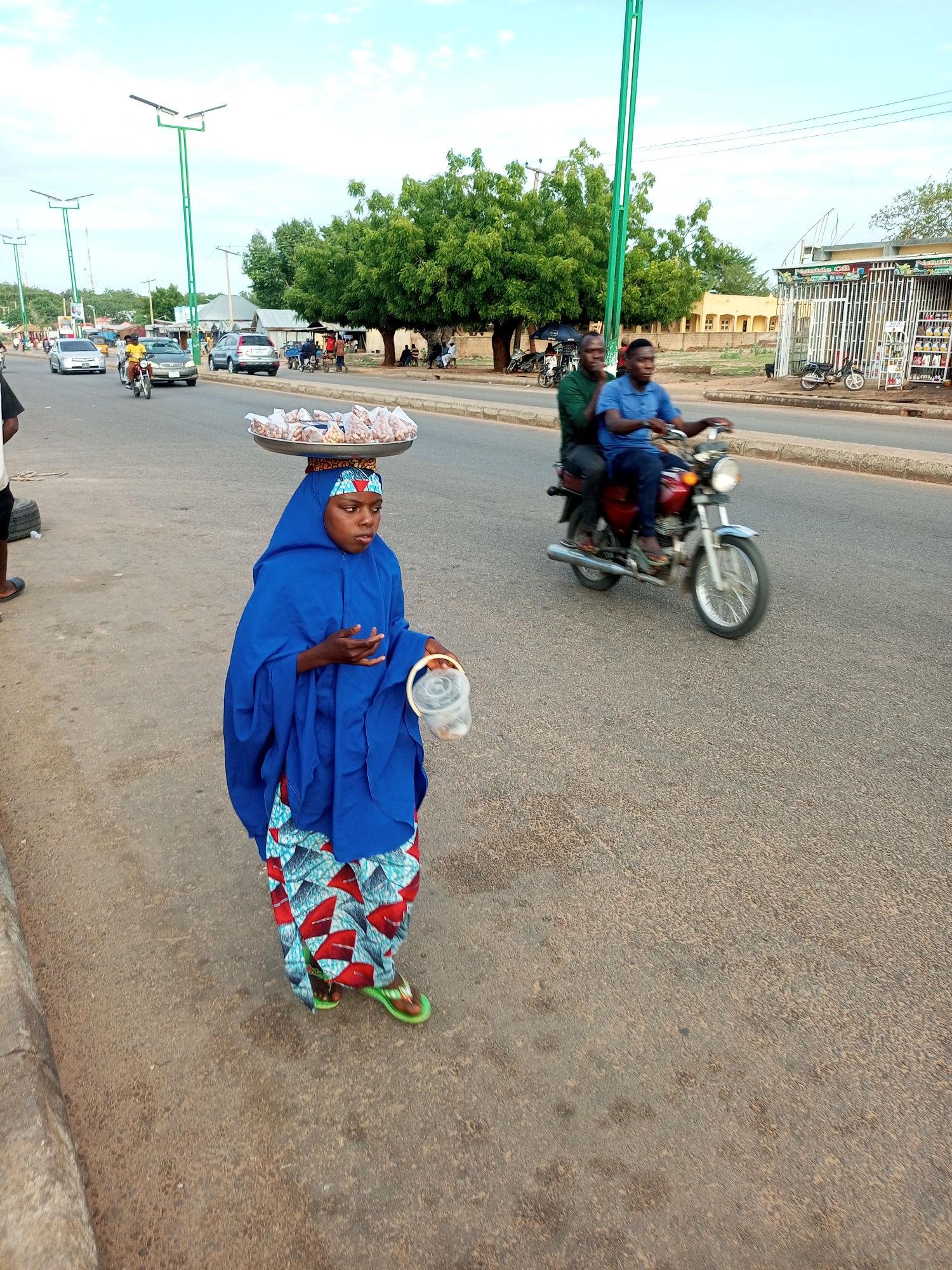 An outdoor street scene captured during daylight hours in the city of Unknown, Unknown. The primary subject, positioned in the foreground-left, is a young woman standing on the shoulder of an asphalt road. She wears a royal blue hijab that covers her head and shoulders, revealing a colorful, patterned skirt or wrapper in shades of red, blue, and white with geometric motifs. Her feet are clad in lime green flip-flops. She balances a large, circular, silver-colored metal tray on her head, which holds numerous small, clear plastic bags, each filled with light brown, oval-shaped items, likely nuts. Her left hand holds a clear plastic container with a white rim and handle, while her right arm is bent at the elbow. She is looking towards the right side of the frame.
On the two-lane asphalt road, positioned in the mid-ground-right, a red motorcycle with chrome accents is in motion, traveling from left to right. Two men ride the motorcycle; the driver wears a blue short-sleeved shirt and dark trousers, and the passenger, seated behind him, wears a dark green short-sleeved shirt and dark trousers. Their faces and the motorcycle's wheels exhibit slight motion blur.
In the background, on both sides of the road, several tall, green streetlights with solar panels are visible. A silver sedan and another light-colored car, along with another motorcycle with a yellow-shirted rider, are present in the distant left lane. To the right, a building with a metal-grated facade, possibly a shop with items displayed on shelves outside, stands beneath a large, leafy green tree. Further back, a light yellow building is discernible. A group of individuals is gathered under a tree on the right side of the road. The road surface is dark grey, textured, and shows signs of wear. A black tire is partially visible on the ground in the far left foreground. The sky is light blue with sparse white clouds.