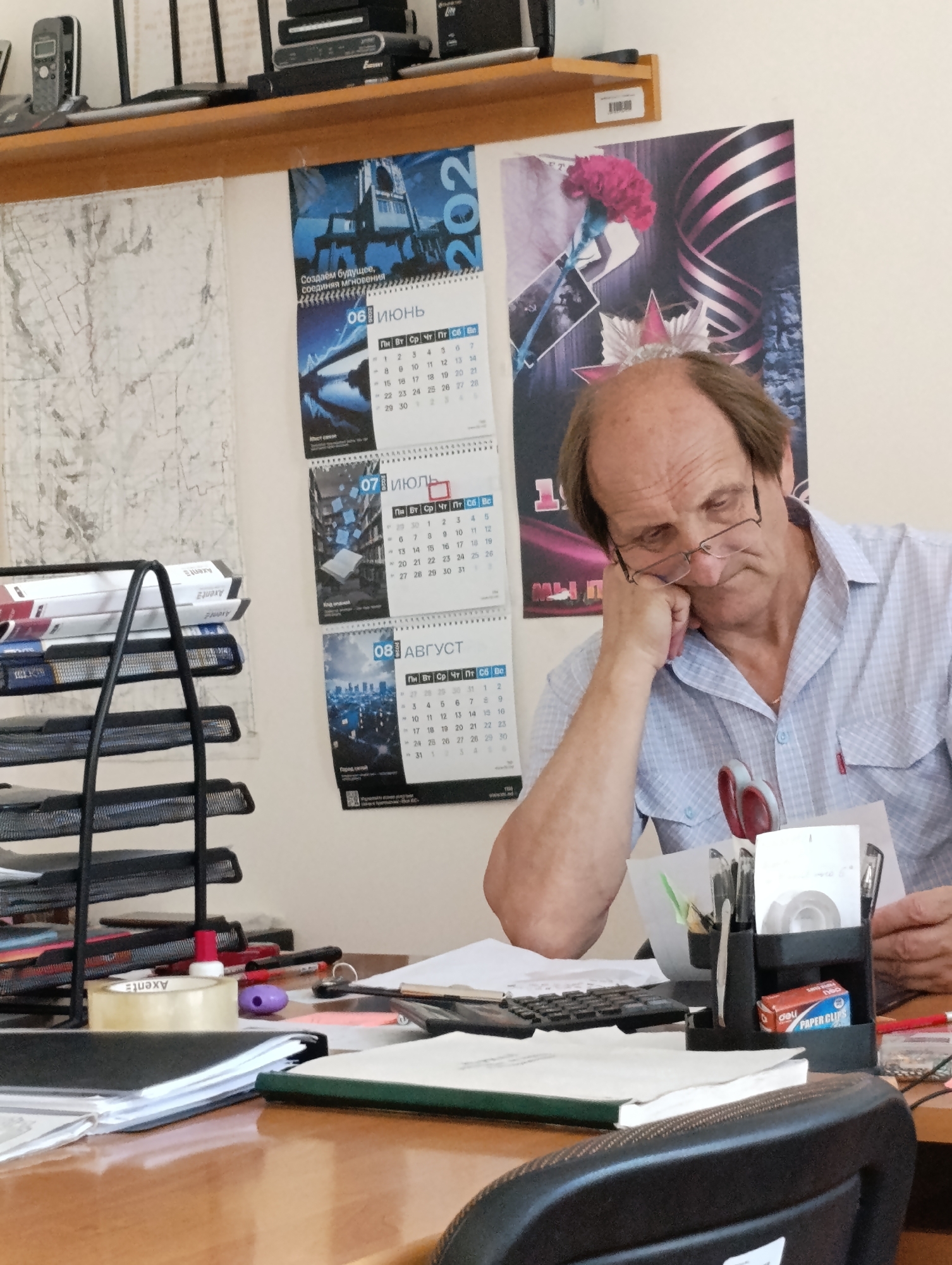 In Dubăsari town council, Moldova, a middle-aged man with thinning brown hair and glasses is sitting at a wooden desk, leaning his head on his hand. He is wearing a blue and white checkered shirt. The desk is cluttered with various office supplies, including stacks of paper, a stapler, pens, a calculator, and a roll of tape. Calendars for June, July, and August of 2020 are visible on the wall behind him, along with a map. A black chair is partially visible in the foreground. The overall environment appears to be an office setting.