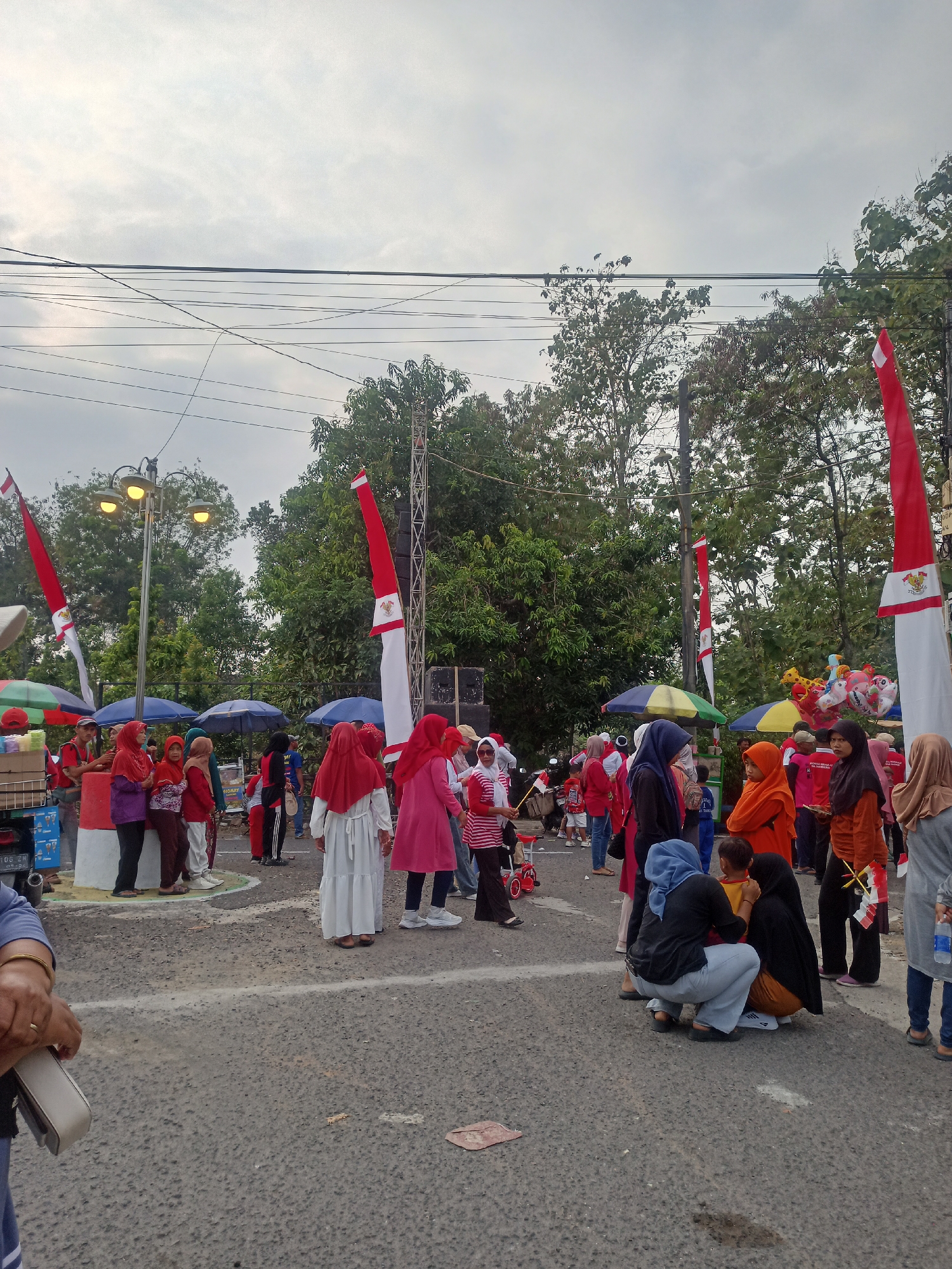 In Karangtalun, Indonesia, a street scene unfolds with a crowd gathered outdoors under a cloudy sky. The atmosphere suggests a local event, possibly a celebration, as evidenced by numerous red and white flags, including some featuring the Indonesian coat of arms, which are prominently displayed. Several individuals are visible, many wearing hijabs and modest clothing in red, white, and earth tones. Some are standing, while others are crouching or sitting on the asphalt ground.
In the background, trees line the perimeter, and utility poles with wires crisscross overhead. Two streetlights are illuminated, casting a warm glow. Several small stalls or carts are present, some with colorful umbrellas, and one displays what appear to be boxes of goods. Balloons, possibly for sale, are visible in the mid-ground. The ground is paved with asphalt, marked with white lines, and shows signs of wear and tear, with some debris present. The overall scene captures a moment of community gathering in an outdoor urban setting.
