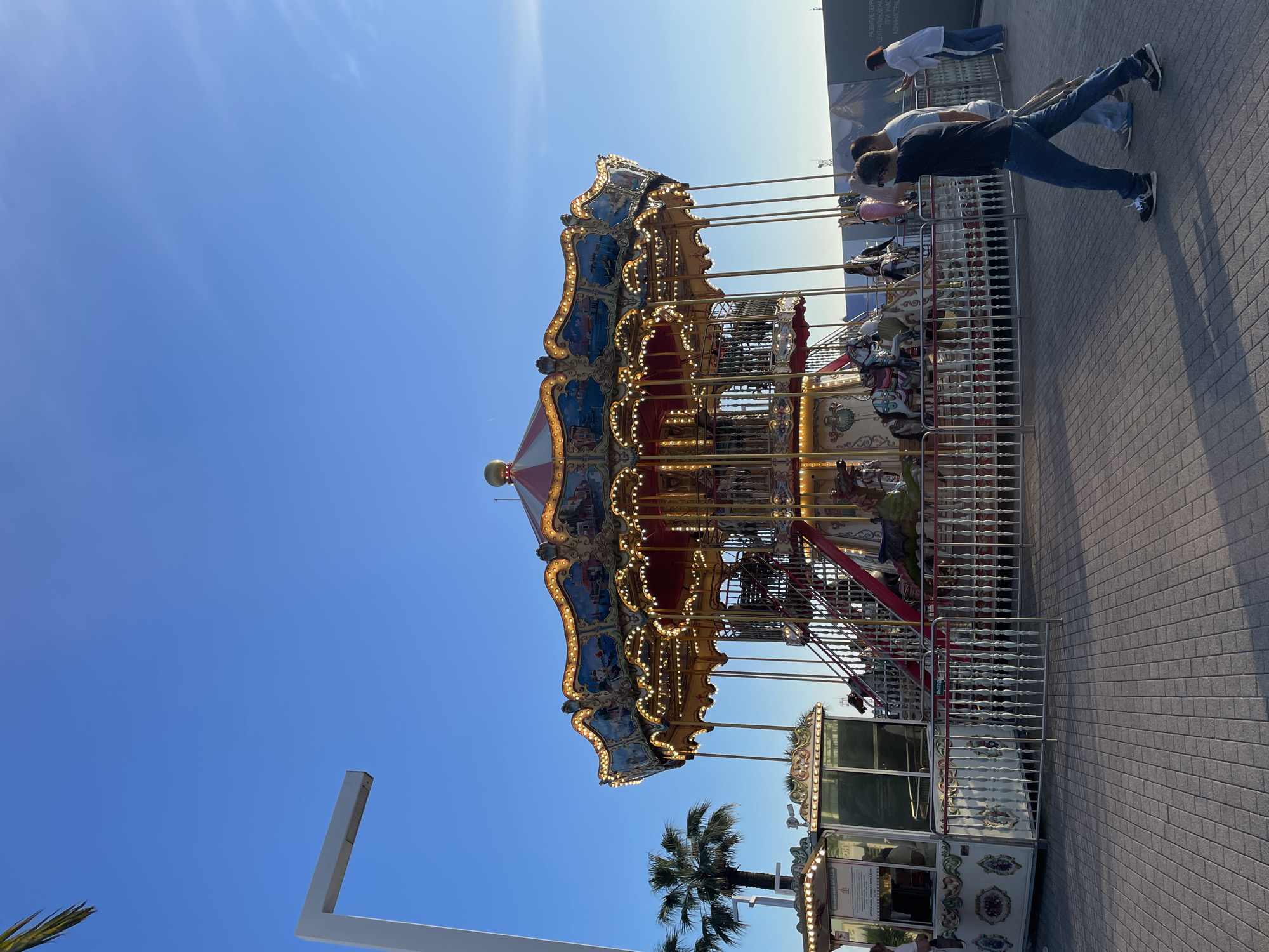 This is a photograph of a carousel situated on a paved promenade in Sochi, Russia. The carousel is ornately decorated with lights and features various rideable figures. In the foreground, three individuals are walking on the promenade, casting shadows on the ground. To the left of the carousel, a ticket booth is visible. Palm trees are present on the left side of the image, and a tall, modern lamppost stands to the far left. The sky is clear and blue. In the background, a construction barrier with imagery is present.