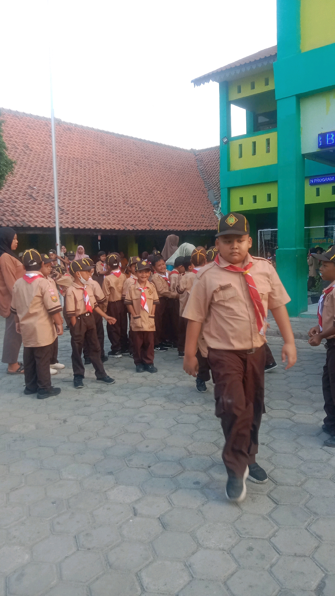 In the city of Jalenjaya, Indonesia, a group of children, likely students, are gathered in what appears to be an outdoor area with hexagonal paving stones. The children are dressed in scout uniforms, consisting of tan shirts, brown pants, and hats, with red and white neckerchiefs. Some of the children are looking at the camera, while others are interacting with each other. In the background, there is a building with a red-tiled roof and a brightly colored building, painted green and yellow, with windows and what looks like a banner or sign on it. The overall scene suggests a school or community event involving young scouts.