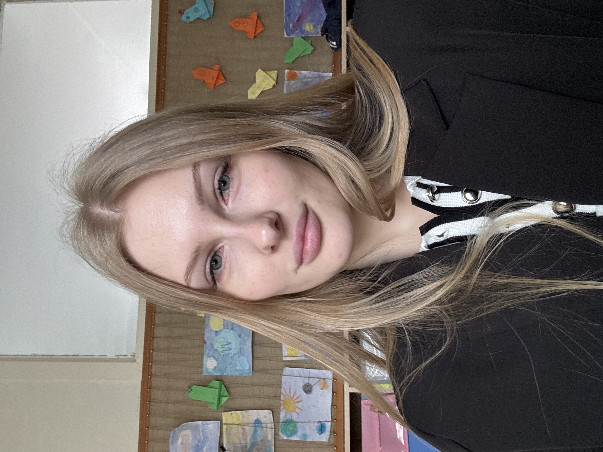 A young woman with fair skin and blonde hair is positioned in the foreground of the image. She is wearing a black jacket over a black and white patterned top. Behind her is a bulletin board displaying colorful children's drawings and origami. The room appears to be a classroom or educational space. There is a white wall or window frame to the left of the bulletin board. The location is Perm, Russia.