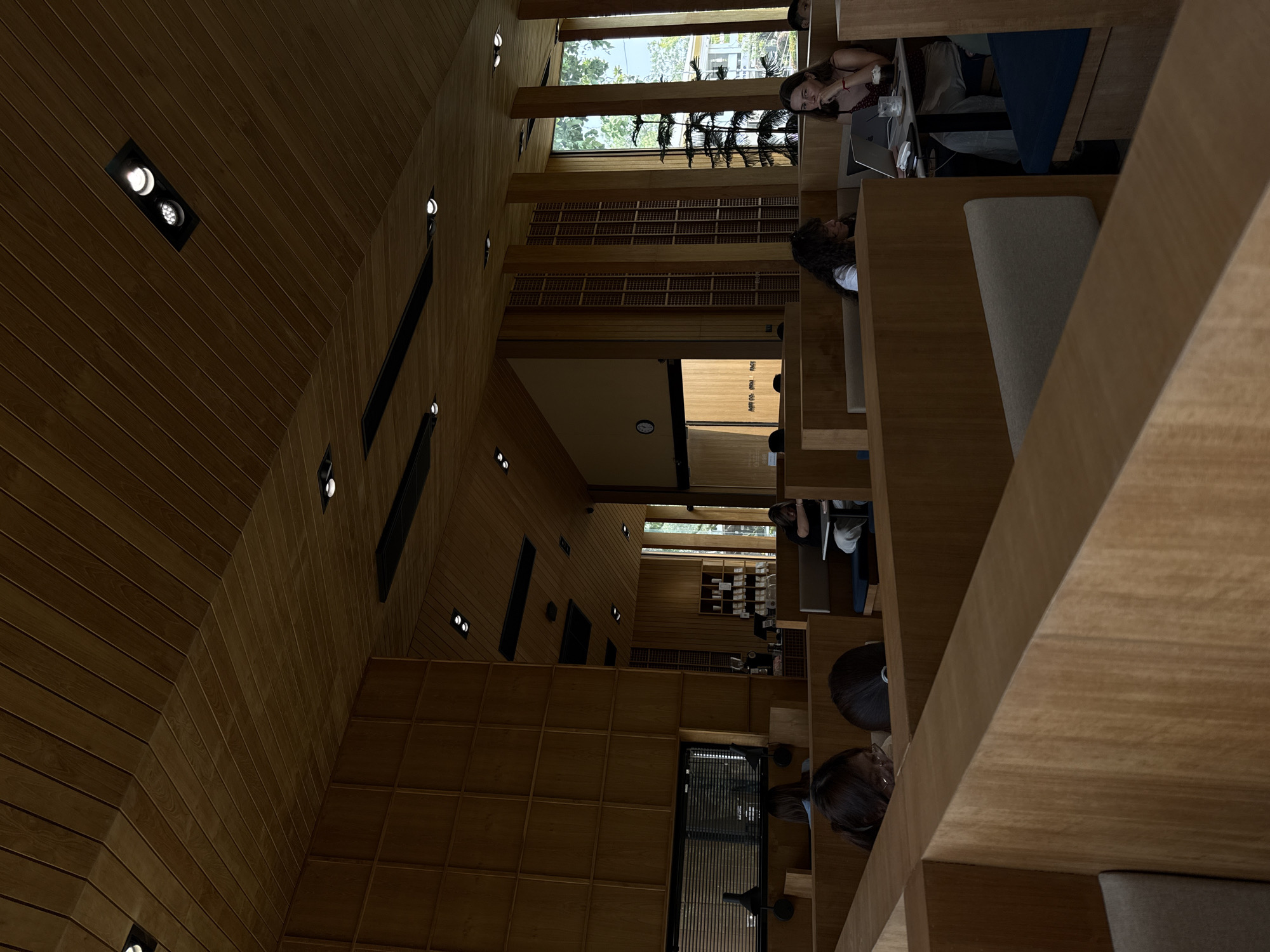 The image depicts the interior of a multi-tiered modern building, likely a library, study lounge, or co-working space, characterized by extensive use of light-colored wooden panels. The walls and ceilings are paneled, creating a warm, contemporary aesthetic with dynamic angles and recessed lighting. Large windows allow natural light to filter in, revealing green foliage and tree branches outside, indicating a daytime setting.
Several individuals are present, mostly seated at individual wooden desks or tables. In the top right, a woman with dark hair sits at a table with a laptop, looking towards the camera. Another person is partially visible next to her. Further down, another woman with curly dark hair is seen from behind, also engaged with a laptop at a similar wooden setup. Other people are scattered throughout the different levels, generally focused on their activities.
The primary activity taking place is quiet work or study, with many individuals using laptops. The space features integrated wooden bookshelves or storage cubbies along the walls. Artificial spotlights are visible in the ceiling and walls, providing additional illumination. The overall atmosphere appears calm and conducive to concentration. No clear, legible text is visible within the frame. The setting is within Chiang Mai City Municipality, Thailand.