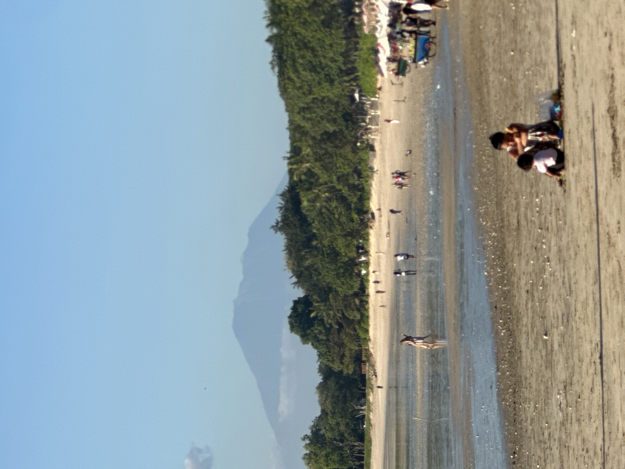 The image depicts a beach scene in Kuta Selatan, Indonesia. In the foreground, two individuals, possibly a mother and child, are seated on the sand. The child is wearing a pink shirt and black shorts, while the adult is wearing a patterned dress and has their arm around the child. A small collection of colorful objects is scattered near them on the sand.
The middle ground shows a shallow body of water extending from the ocean onto the sandy beach. Several people are visible walking in the water or on the wet sand, including a woman in a long dress walking with another person. Further back, on the dry sand, there are more people, beach umbrellas, and what appears to be a food cart with people gathered around it. A line of lush green trees forms the backdrop to the beach.
In the distance, a large, imposing mountain dominates the horizon under a clear blue sky. The mountain appears to be dormant, with a soft, hazy outline. The overall impression is a serene, daytime scene at a popular Indonesian beach.