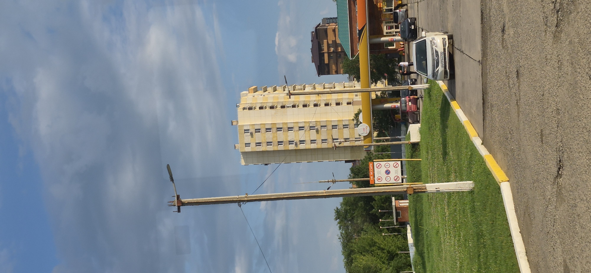 The image depicts a street scene in Ussuriysk, Russia, focusing on a gas station and a tall residential building. The sky is blue with scattered clouds, indicating daytime. In the foreground, a paved road with a yellow and white striped curb borders a grassy area. A lamppost stands to the left, with wires extending across the scene.
The gas station, identified by a yellow canopy and the "НК УССУРИ" logo, is a prominent feature. Several cars are visible at the gas station, including a white sedan in the foreground, a red hatchback, and darker sedans further back. Two individuals are standing near the gas pumps, interacting with each other.
Behind the gas station, a multi-story yellow residential building with numerous windows stands tall. To the right of this building, another, shorter, darker-colored building is partially visible. Trees and some smaller structures, possibly utility buildings, are situated behind the grassy area to the left. A sign with multiple circular symbols is posted near the lamppost. The overall impression is an urban setting with a mix of commercial and residential elements under a bright sky.