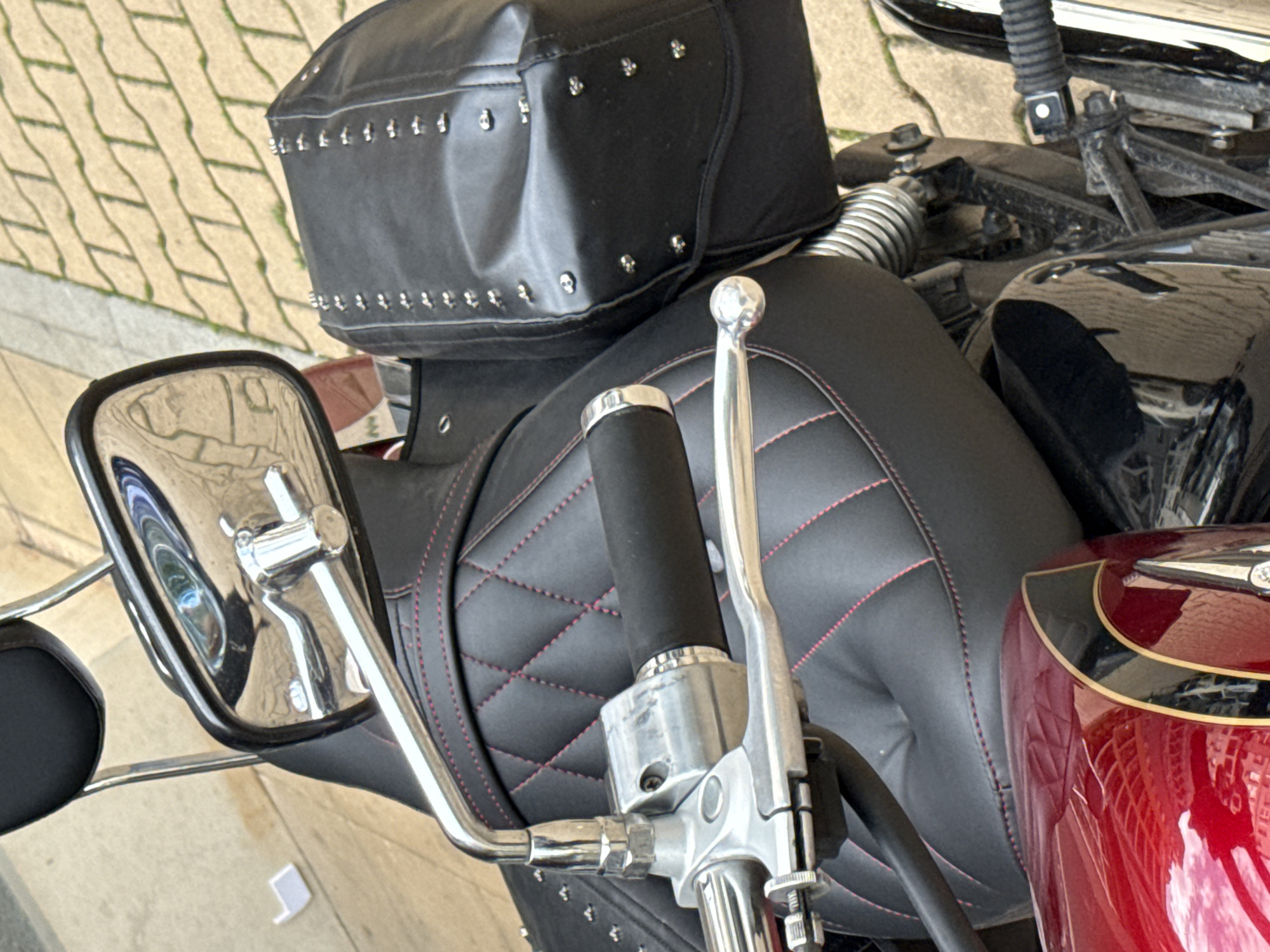 A red and black motorcycle is parked outdoors on light-colored paving stones in Sofia, Bulgaria. The motorcycle features a glossy, deep red fuel tank with black and gold pinstriping, a black quilted seat detailed with red stitching, and a black studded saddlebag affixed to the rear. Chrome elements include the right rearview mirror, handlebars, control levers, and a visible suspension spring coil. The right handlebar is equipped with a black rubber grip adjacent to a silver metallic control housing and brake lever. The chrome mirror reflects portions of the ground and a light-toned background. The ground is composed of rectangular, light-colored pavers laid in a pattern, upon which a small, white, folded paper object rests to the left of the motorcycle. No individuals are visible.