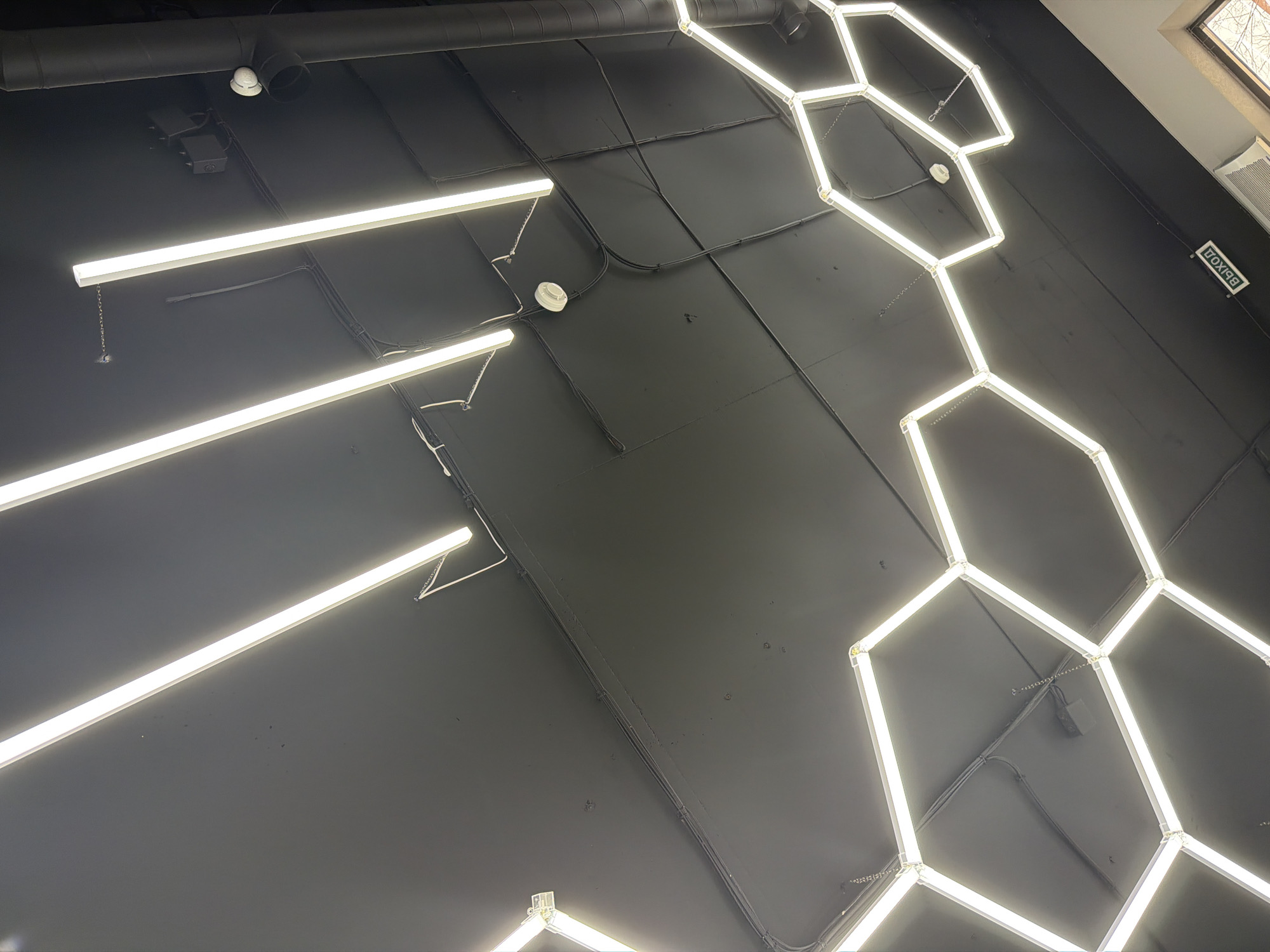 The image shows a ceiling with installed lighting. On the left side of the image are three parallel, illuminated linear light fixtures. Toward the right side of the image are several interconnected hexagonal light fixtures. The ceiling surface is dark and has black conduit, wires, and fixtures attached. At the top left of the image is black ductwork. At the top right of the image is a window and an exit sign. The location is unknown, however, it is not specified the city is Krasnoyarsk, Russia.