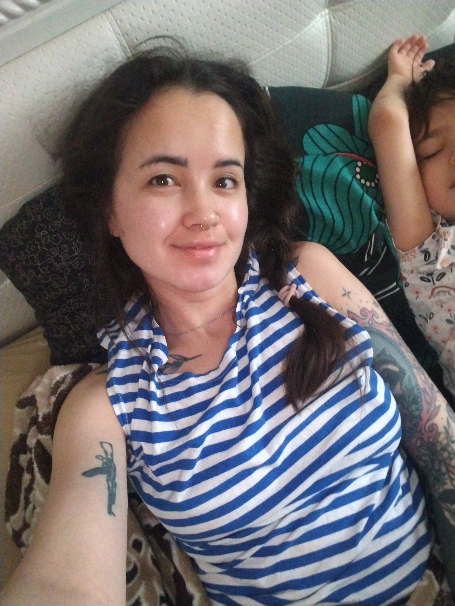 This media file captures a selfie of a woman lying down next to a sleeping child, likely indoors in Noviy, Russia. The woman, positioned in the foreground, has dark hair, pulled into a braid on her right side secured with a pink elastic. She has light brown eyes and a faint smile, looking directly at the camera. A small gold-colored hoop nose ring is visible on her right nostril. She wears a blue and white horizontally striped sleeveless top.
The woman exhibits several tattoos: a dark, abstract design on her left upper arm; a large, colorful sleeve tattoo on her right arm, featuring intricate floral or nature-themed patterns in blues, greens, and possibly other hues; a small, dark tattoo resembling a bird or abstract shape on her chest/collarbone area; and a small star-like tattoo on her right upper arm.
Beside her, a child with dark hair is asleep, their face partially obscured and one arm resting above their head. The child is wearing a light-colored top with a subtle, possibly floral pattern. They are lying on a pillow with a teal and green abstract or floral design.
The setting appears to be a bedroom, with a white, textured headboard visible in the background. Other pillows with dark floral or paisley patterns are also present. The soft lighting suggests it is daytime. The overall impression of the image is one of calm and domestic intimacy. There is no visible text.