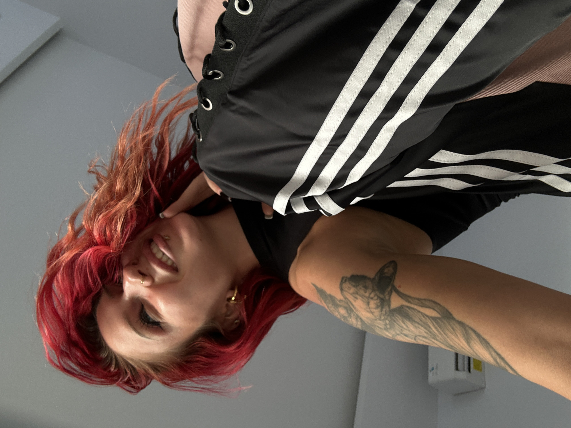In this low-angle shot, a woman with vibrant red hair and piercings in her nose and lip smiles upwards. She is wearing a black sleeveless top, and her left arm, adorned with a tattoo of a bird, is visible. The woman is also wearing black athletic pants with white stripes down the side, featuring metal eyelets and a lace-up detail at the waist. A pink fabric is visible underneath the pants. The background appears to be an indoor setting with a plain wall and a white air conditioning unit. The scene does not provide any specific indicators of its location within North Kuta, Indonesia.