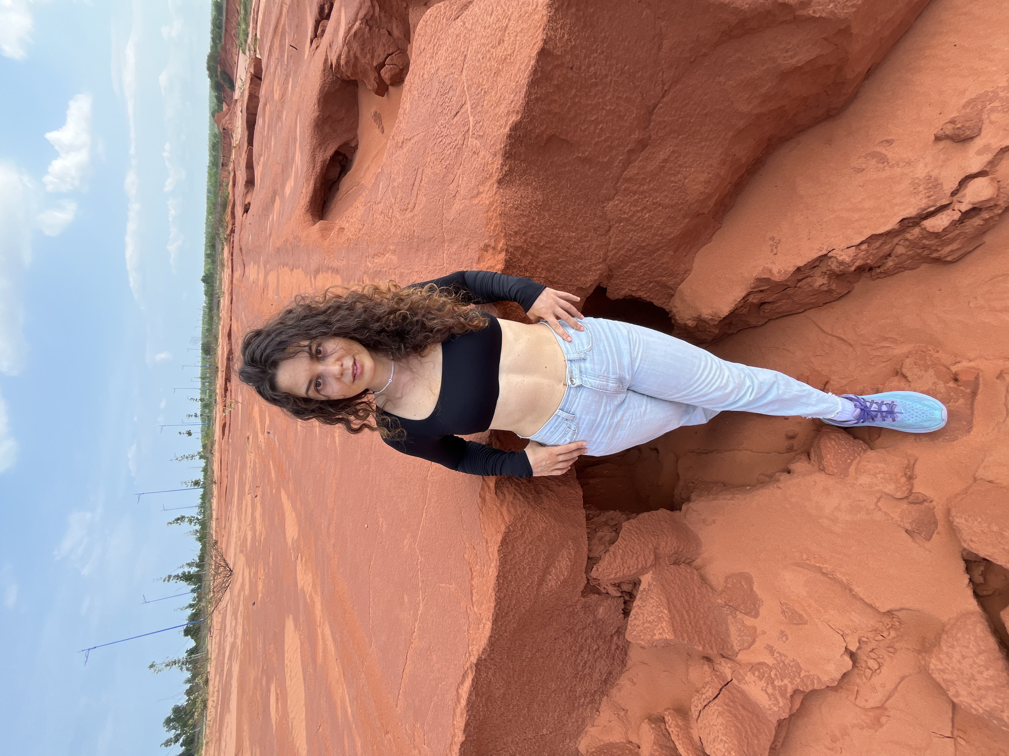 A young woman with curly dark hair stands in a landscape of eroded red rock formations under a cloudy blue sky. She is wearing a black long-sleeved crop top, light blue jeans, and blue and purple athletic shoes. Her hands are placed on her hips. The ground is covered in red sand and rock, with some small green shrubs and tall, thin light poles visible in the background. This landscape is characteristic of the Mui Ne Sand Dunes in Vietnam.