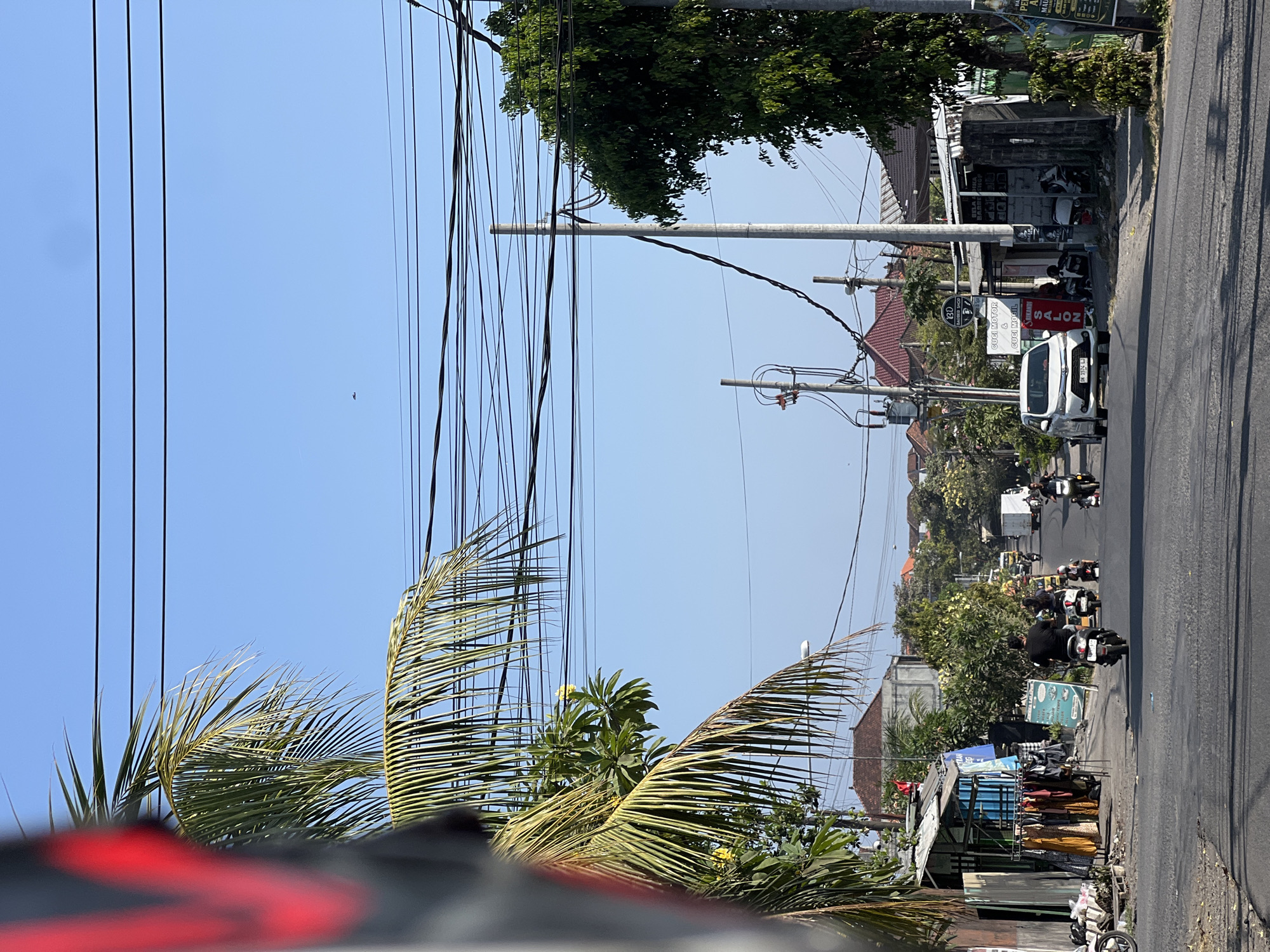 This image depicts a street scene in Denpasar, Indonesia, on a clear, sunny day. The foreground is dominated by the road, with a white car and several motorbikes in motion. On the left side of the road, there are small shops selling clothing displayed on racks. Further down the street, there are more shops, including a barber shop. Palm trees and other foliage are visible along the sides of the road, and utility poles with numerous overhead wires crisscross the sky. The architecture of the buildings suggests a tropical setting, with tiled roofs and open-air structures. The overall atmosphere is that of a bustling urban environment.