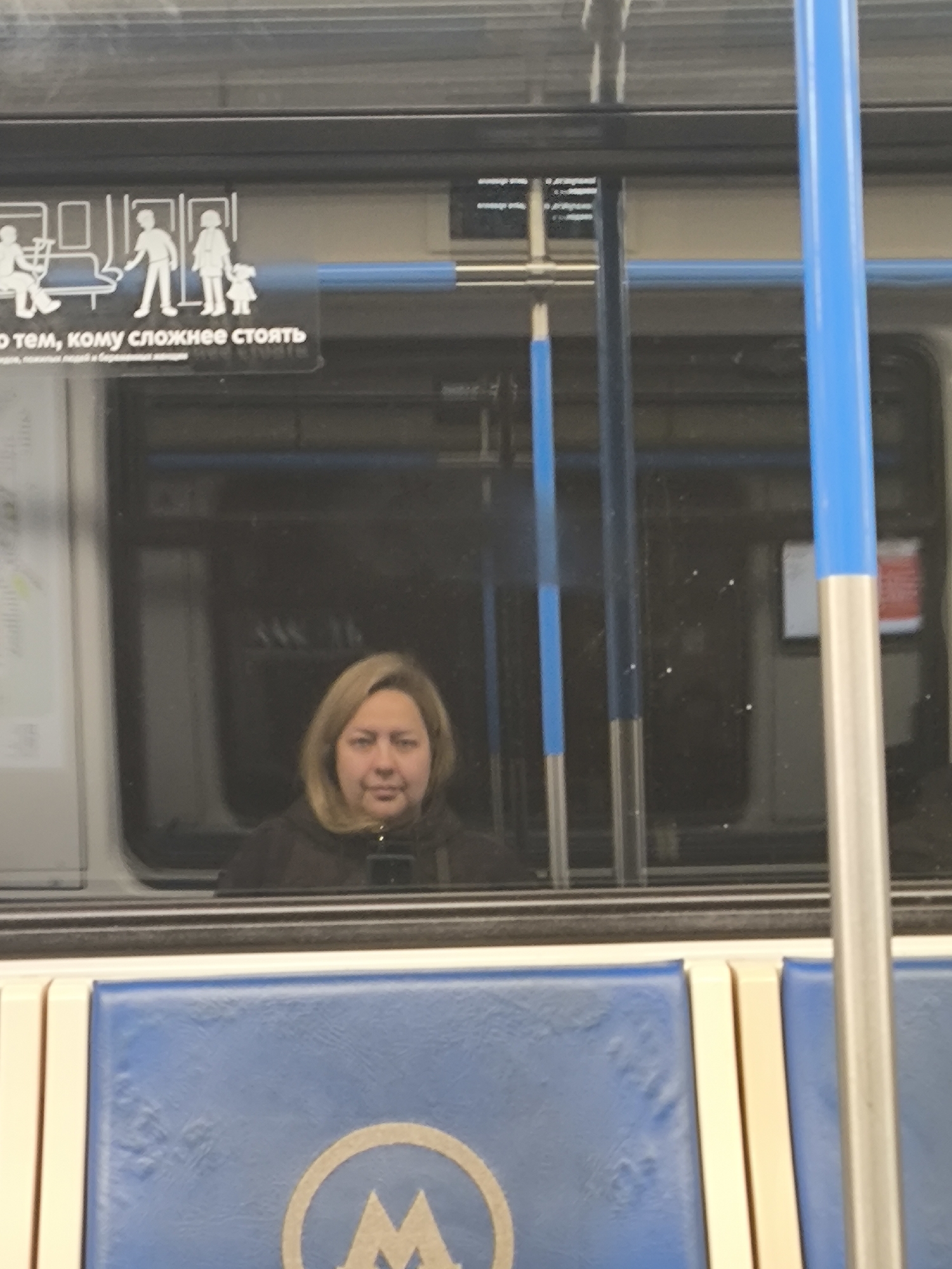 A woman with blonde hair is reflected in a subway car window. The subway car has blue padded seating, a blue grab bar, and signage in Russian above the window with stick figures. The "M" logo for the Moscow Metro is on the seat.