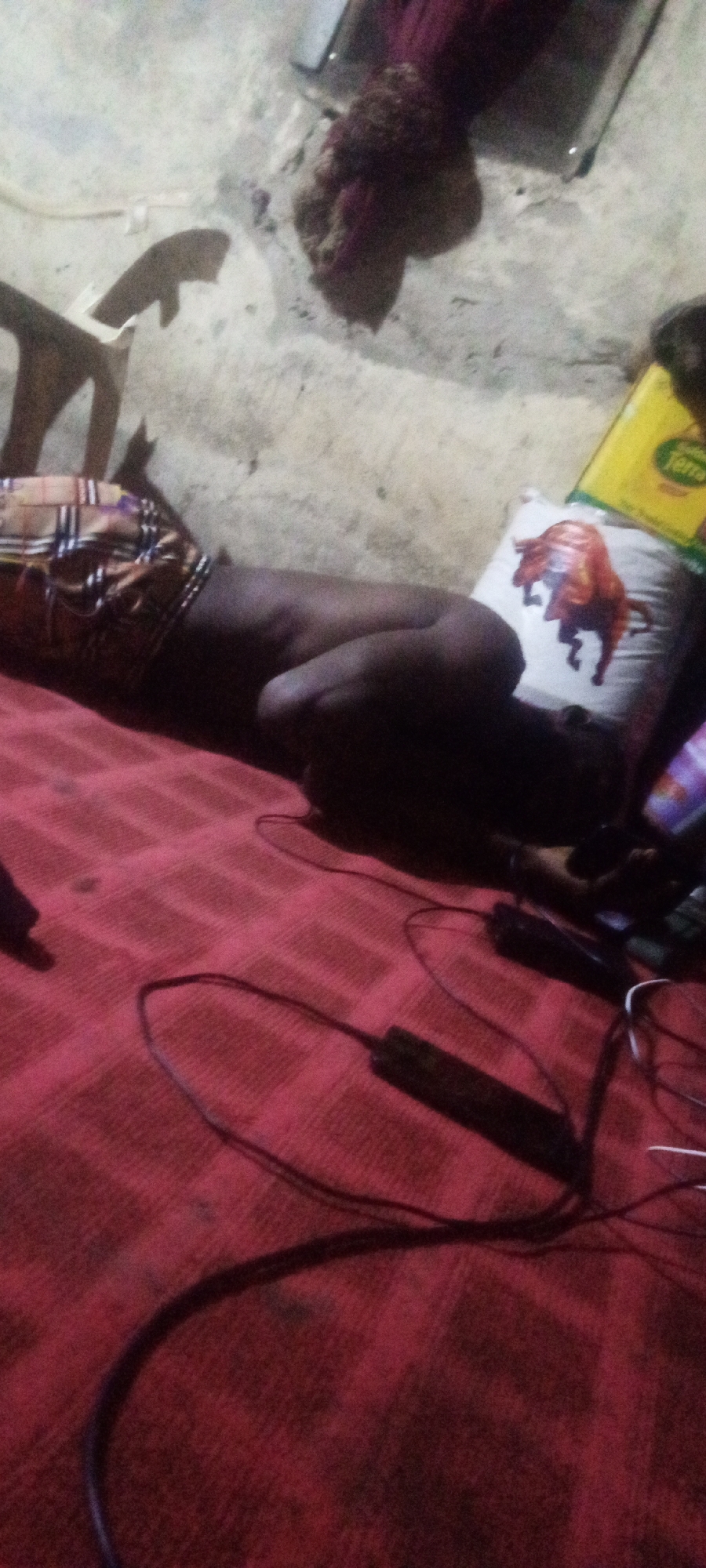 The image captures an indoor scene in Sabon Tasha, Nigeria, featuring a person lying on their side on a red mat with a subtle checkered pattern. The individual, dressed in a plaid or patterned garment around their waist, appears to be asleep or resting, with their face turned away from the camera.
Near the person's head rests a white pillow adorned with a prominent illustration of a brown and orange bull. Adjacent to the pillow, a yellow rectangular container, possibly for food or oil, is visible, with the word "Terra" clearly legible in green lettering. Numerous dark cables and electronic accessories, possibly chargers or power strips, are scattered across the red mat, suggesting devices are connected or in use nearby.
The background reveals a light-colored wall, possibly concrete or mud, showing signs of wear and dark smudges. Portions of indistinct furniture or structures, including what might be a wooden chair, are visible in the upper left corner. The lighting in the room is dim and artificial, suggesting the image was taken at night or in a poorly lit interior space. The overall impression is that of a humble, domestic setting.