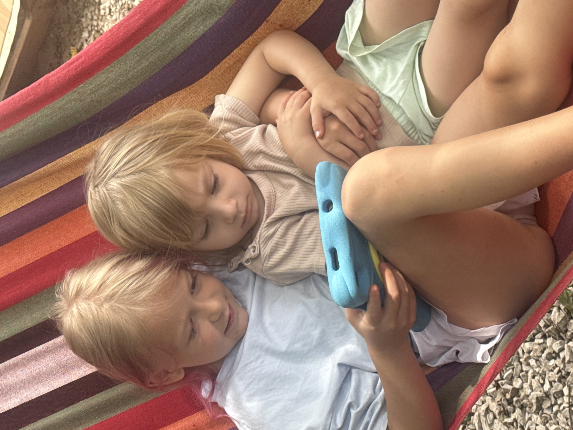 Two young children are lying in a colorful striped hammock, oriented diagonally from the top left to the bottom right of the frame. The hammock features stripes in shades of red, green, purple, orange, and brown.
The child in the upper part of the image has blonde hair and is wearing a light-colored, possibly beige or light brown, ribbed long-sleeved shirt. Their eyes are closed, suggesting they might be sleeping or resting. Their hands are clasped together on their chest.
The child in the lower part of the image also has blonde hair and is wearing a light blue or white short-sleeved top. Their eyes are partially open, and they appear to be looking downwards or slightly to the right with a faint smile. This child is holding a blue and yellow object, possibly a toy or a tablet, with both hands.
The children's legs and lower bodies are partially visible, with one wearing light-colored shorts. The overall lighting suggests it is daytime, likely outdoors, given the presence of small stones or gravel visible at the top left and bottom right corners, outside the hammock. The mood appears peaceful and relaxed. There is no visible text in the image.