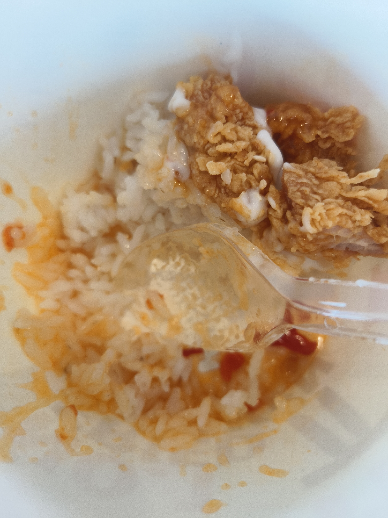 The image shows a close-up view of a bowl of food. The primary subjects are rice and fried chicken pieces, coated in a sauce that appears to be a mixture of sweet and savory flavors, possibly with some chili. A clear plastic spoon is partially submerged in the rice, indicating that someone is in the process of eating. The lighting suggests it's daytime, and the food is presented in a disposable container, hinting at a casual dining or take-out meal. The overall impression is one of a simple, satisfying meal. There is no visible text in the image. Given the location context of Denpasar, Indonesia, this could be a common dish found in local eateries or street food stalls. The food appears to be hot, with some condensation visible on the container.