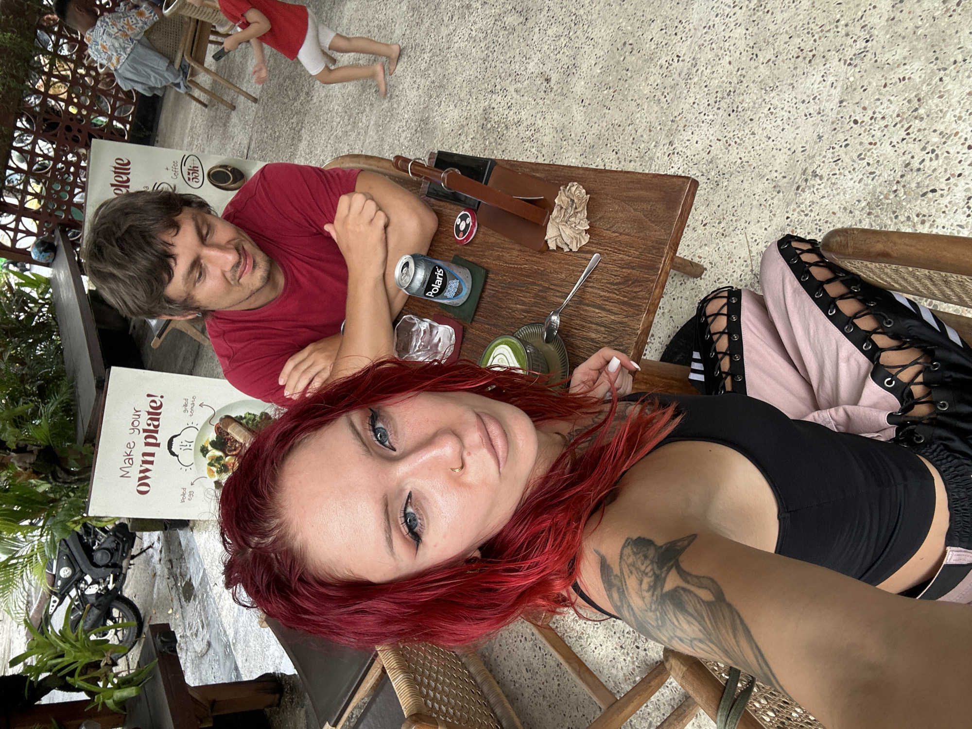 The image depicts a lively outdoor seating area at a cafe in North Kuta, Indonesia. A woman with bright red hair and tattoos on her left arm is in the foreground, posing for the camera. She is wearing a black tank top and pink pants with black lace-up detailing. To her right, a man in a red t-shirt is seated, looking down. A wooden table between them holds a glass of water, a can of "Polaris Soda Water," a small round object with a red and white pattern, and what appears to be a serving set with a brown handle. In the background, a motorcycle is parked near a sign that reads "Make your own plate!". A child in a red shirt and white shorts is visible further in the background, walking away from the camera. Other patrons are seated at tables, and the environment suggests a casual, tropical setting with plants and an open-air layout.