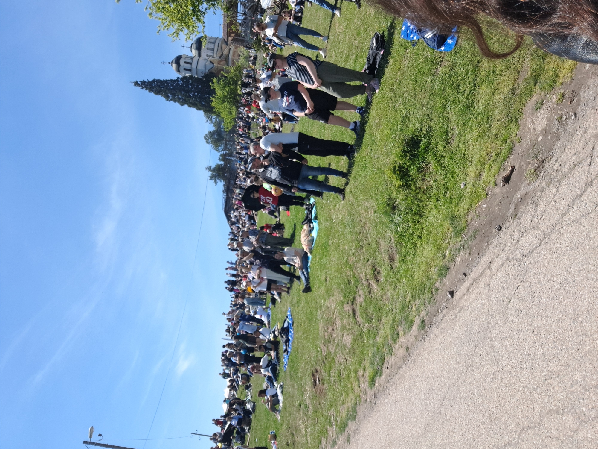 The image shows a large crowd of people gathered on a grassy field next to a road under a blue sky. A church with dark domes is visible in the background. The location is Sofia, Bulgaria. The crowd appears to be watching something, though the specific event or attraction isn't clear. People are standing, sitting, or milling about. A few people are closer to the camera, and appear to be casually dressed. There is a sidewalk or paved area near the grassy field.