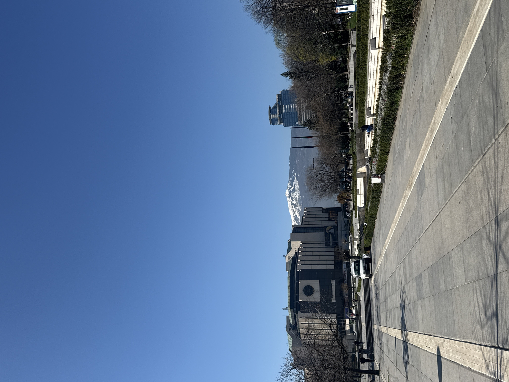 This image shows a wide, paved pedestrian area in Sofia, Bulgaria, likely a central plaza. The sky is clear blue. Buildings line the plaza, including the National Palace of Culture (NDK) and a modern high-rise office building. A snow-capped mountain (likely Vitosha Mountain) is visible in the background. Bare trees are scattered throughout. People and a white van are visible along the edge of the paved area. Hedges and landscaping separate the pavement from the buildings. Shadows from trees are cast on the pavement.