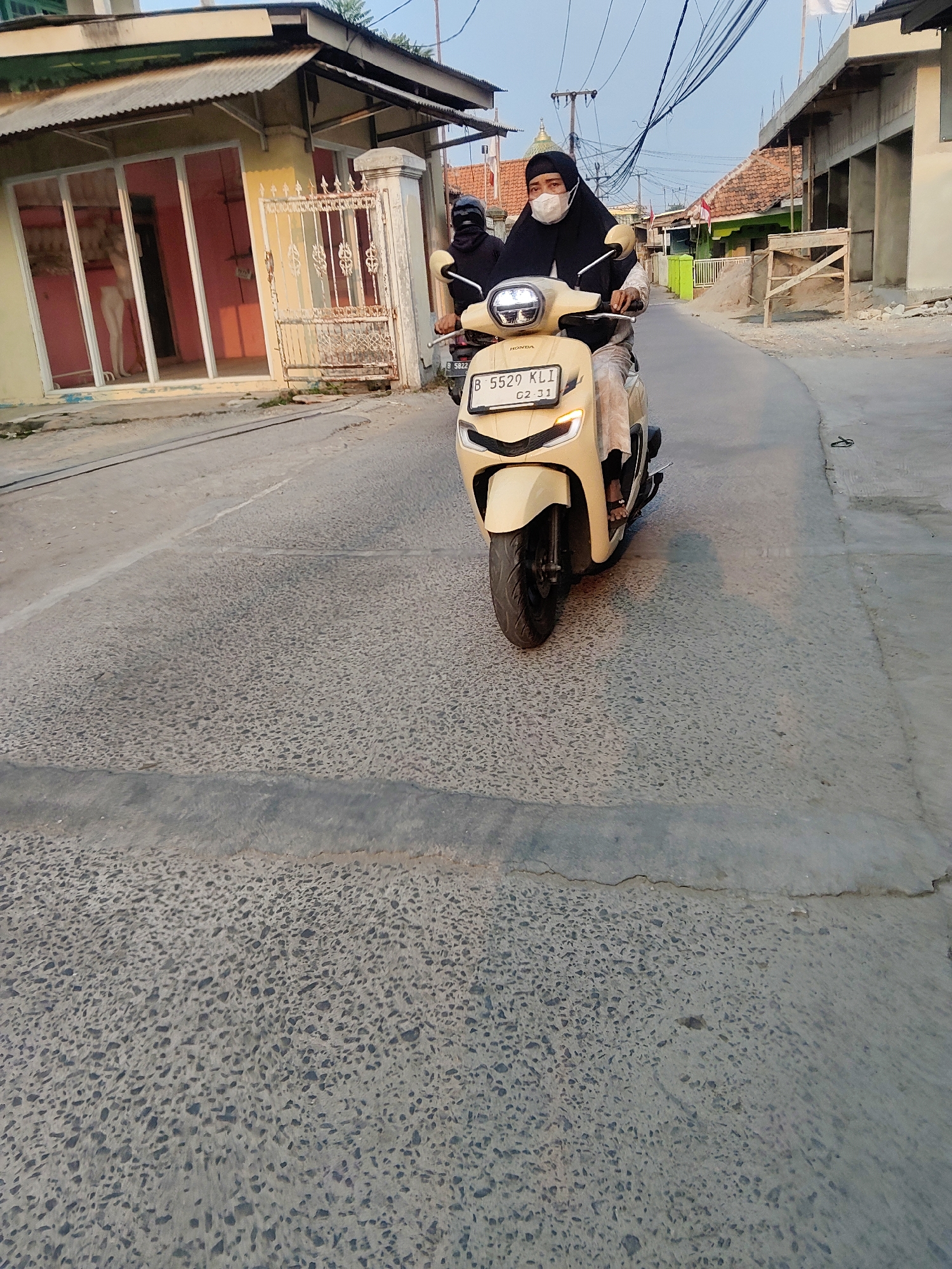 A person wearing a black hijab and a white mask is riding a light beige Honda scooter down a street in Mekarmukti, Indonesia. The scooter has a license plate that reads "B 5529 KLI 02-31". The person is wearing a black jacket and light-colored pants. In the background, there are buildings with corrugated metal roofs and some construction materials. The street is paved with asphalt. The sky is clear and there are power lines overhead. A mannequin is visible in the window of a building on the left.
