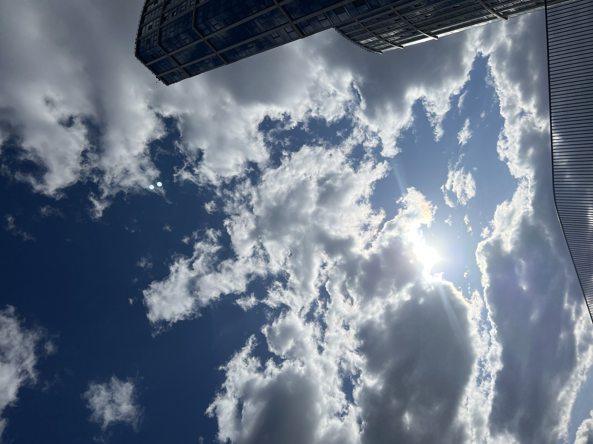 The image displays an upward view of a dynamic sky, framed by modern architectural structures, set in an urban environment, likely in Khimki, Russia.
The sky is a deep blue, partially obscured by numerous clouds. The clouds vary in appearance, ranging from bright white, fluffy cumulus formations to denser, darker grey clouds, suggesting a partly cloudy or dramatic weather pattern. In the upper right section of the frame, the sun is intensely bright, peeking through the clouds and creating a prominent sunburst effect with visible rays of light. Several circular light artifacts, possibly lens flares or reflections, are present, including two distinct white orbs in the mid-left blue sky and another near the sun.
Two tall buildings are visible at the top edges of the frame. The building on the upper left features a modern, glass-clad facade with a grid pattern of windows, reflecting the sky and clouds. The building on the upper right has a distinctive, slightly curved structure with a facade composed of fine horizontal lines or slats, giving it a ribbed or patterned texture.
The scene captures a bright, daytime setting. No specific activities or individuals are depicted, and no visible text is present.