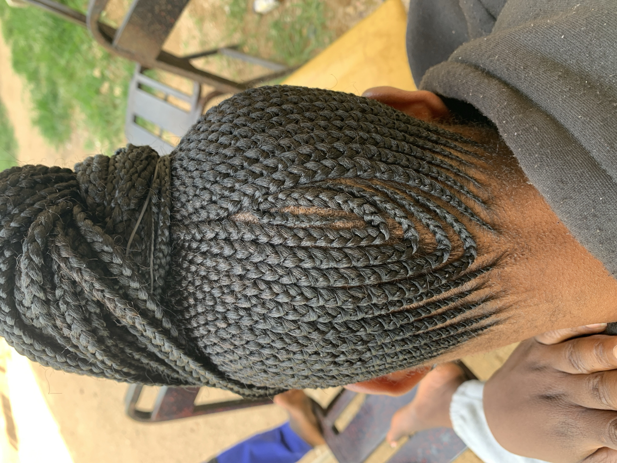 In Jos, Nigeria, an individual's head is shown from behind, focusing on a tightly braided hairstyle. The braids are dark and meticulously styled, forming a large bun at the crown and fanning out in a V-shape down the back of the head. A clear parting is visible down the center of the scalp, with smaller braids creating a textured pattern. The person is wearing a dark hooded jacket. In the background, out-of-focus elements suggest an outdoor setting with glimpses of furniture and greenery, consistent with a residential or recreational area in the city.