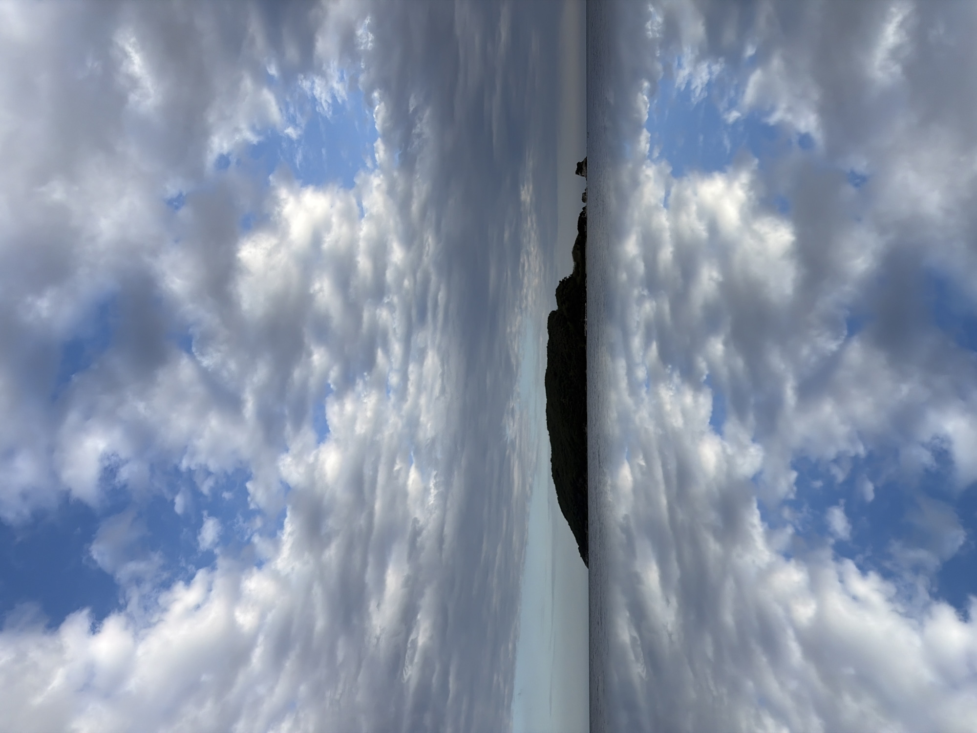 The image is a mirrored scene of clouds and sky, with a landmass or island visible in the center. The sky is mostly blue with patches of white and gray clouds. The landmass is dark and appears to be covered in vegetation, likely a rocky coastline or island. Given the location of Budva, Montenegro, it is possible this is a view of Sveti Nikola Island from the coast. The mirrored effect emphasizes the clouds and creates a symmetrical composition.