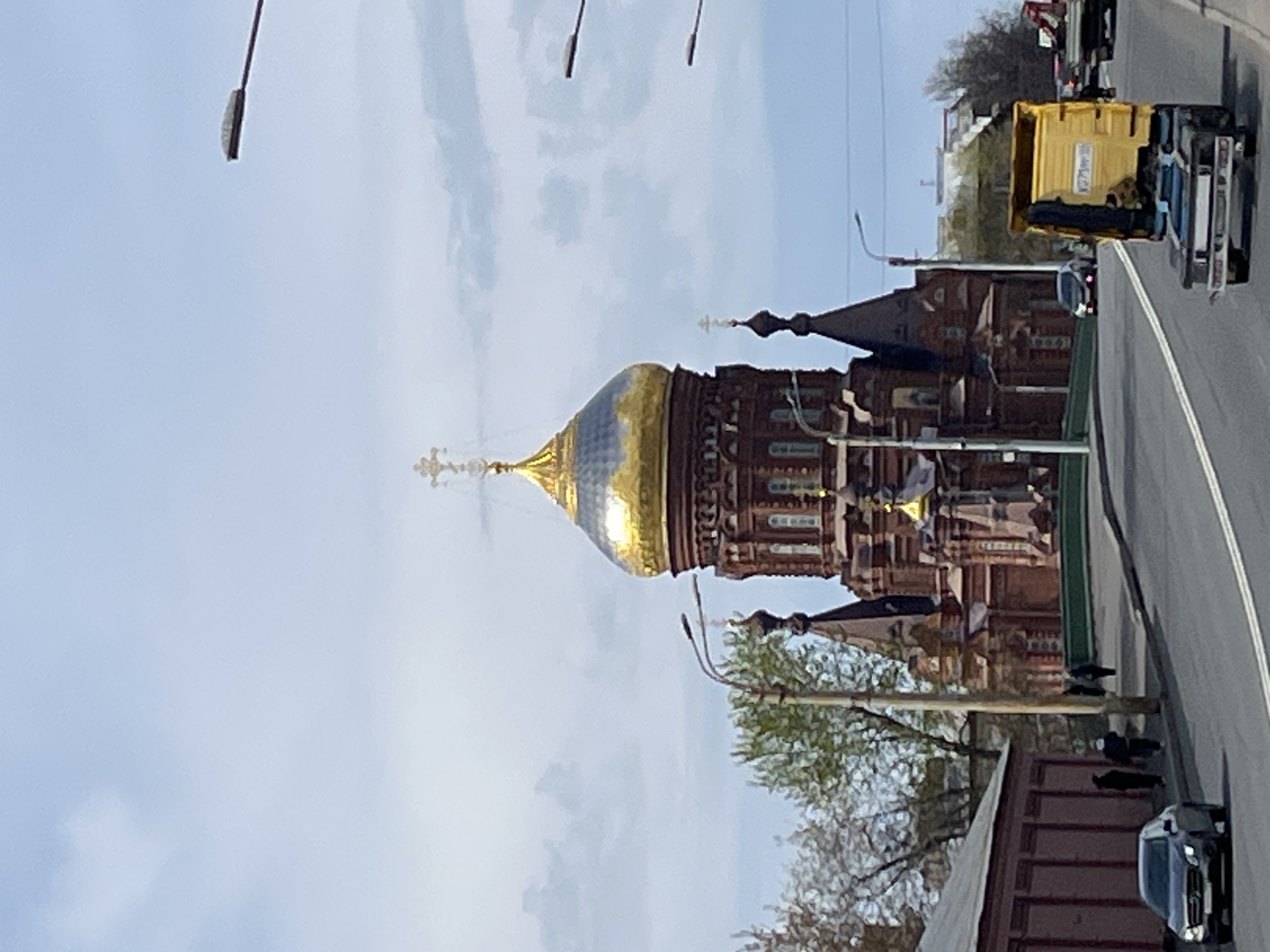 The image shows a red brick Orthodox church with a large golden dome and cross, indicative of Russian religious architecture. This is likely the Church of the Savior on Blood in St. Petersburg, Russia, given the architectural style. A road runs along the right side of the image, with several vehicles, including a box truck. A pedestrian may be visible on the sidewalk. Streetlights are seen on the side of the road. The sky is overcast with light blue and white clouds.