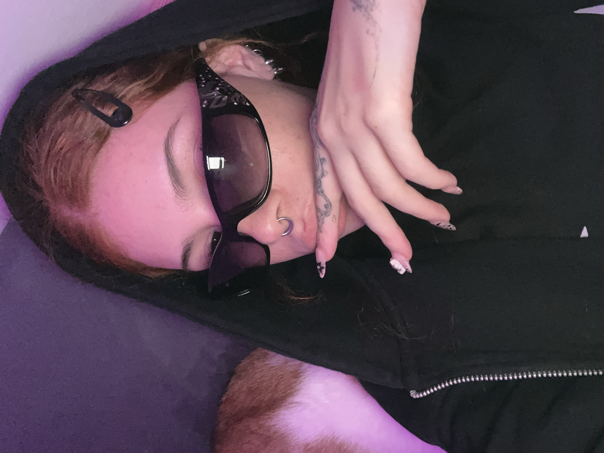 A person lies on a purple surface wearing a black hooded sweatshirt with a visible zipper. The person is angled towards the left and is illuminated with purple lighting. They wear oversized sunglasses, a nose ring, and multiple earrings. A black hair clip is visible in their auburn hair. Their right hand is raised with fingernails painted with a black and white pattern, and a tattoo is visible on their hand and wrist. Part of a brown and white animal, possibly a pet, is visible in the lower left corner. The location is unknown, and there is no indication it is in Buenos Aires, Argentina.