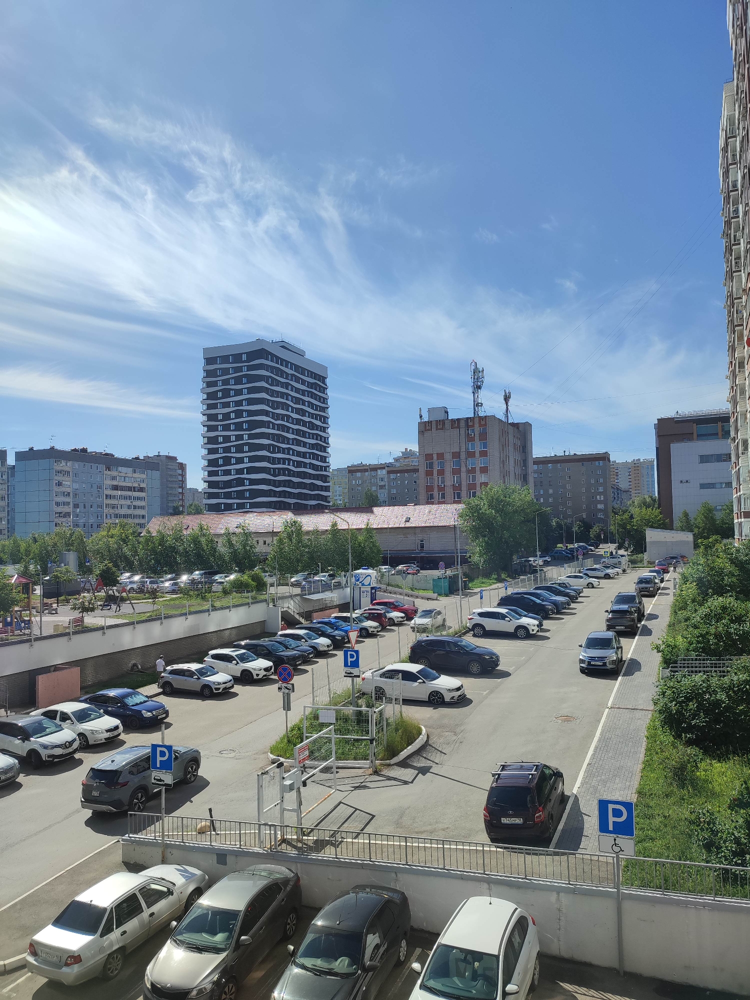 The image shows an outdoor scene with a parking lot, buildings, and a playground. In the foreground, several cars are parked. A gray SUV is parked in the lower right, with a "P" sign indicating parking for disabled individuals. To the left, a white hatchback is parked. Further back, a line of parked cars stretches across the lot.
In the midground, a playground with swings and slides is visible. Beyond the playground, a modern, multi-story building with a wavy, dark facade stands out. To its left, older, taller residential buildings are visible. A cellular tower is located on top of one of the buildings in the background.
The environment is a sunny day with a blue sky and scattered white clouds. Trees and green foliage are present throughout the scene, especially on the right side of the parking lot. A few individuals are sparsely scattered around the area. The scene appears to be a residential or mixed-use urban area.