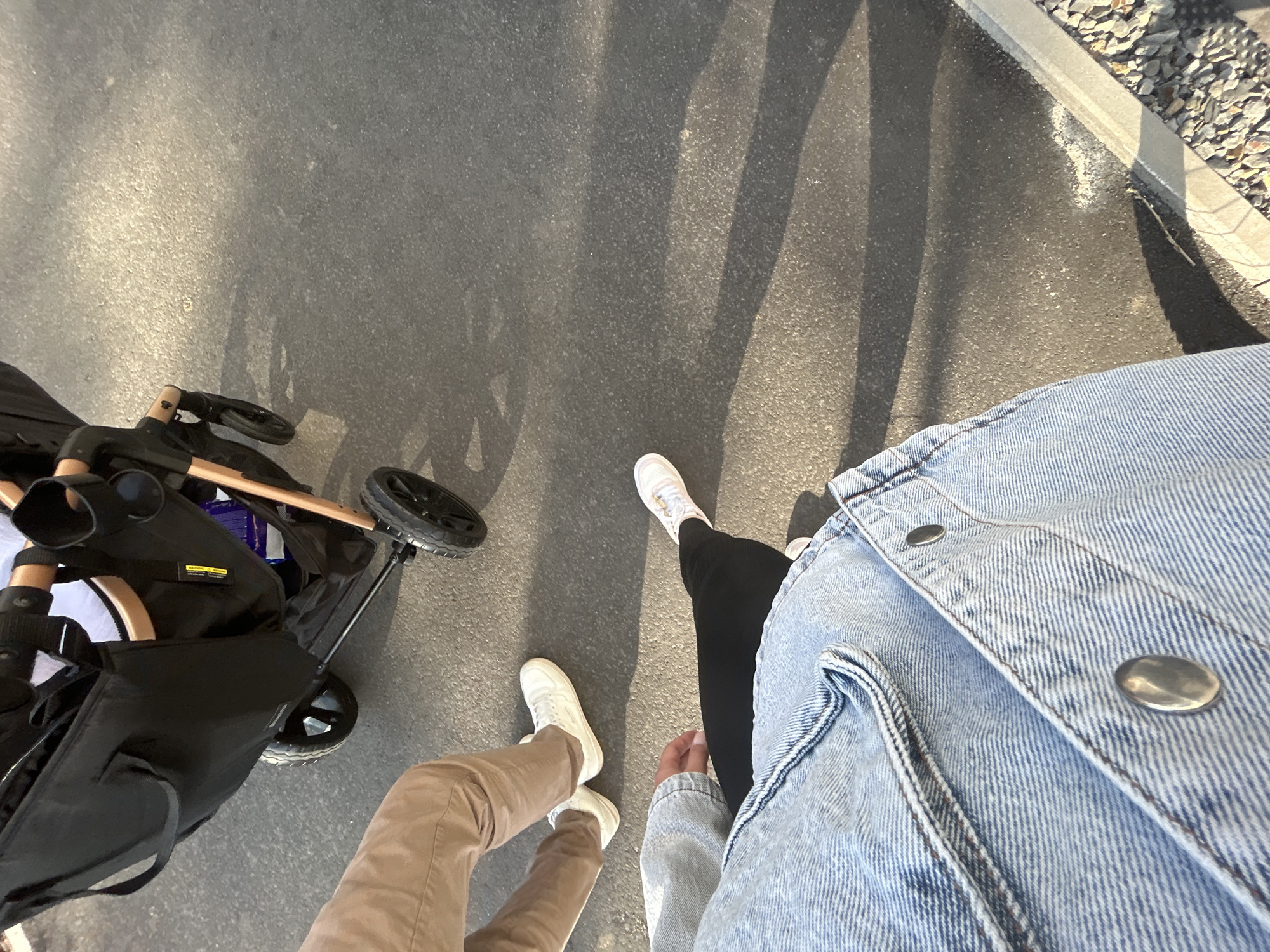 Two individuals are walking on an asphalt surface. The person in the foreground is wearing a light blue denim jacket and black leggings, with one foot visible, wearing a white sneaker with pink accents. They are in the process of walking forward. Behind them, another person is walking and pushing a black stroller. This individual is wearing tan pants and white sneakers. The stroller has a wooden handle and black wheels. The ground is a dark asphalt surface, with shadows cast by the stroller and possibly other unseen objects. In the lower right corner, a concrete curb is visible, adjacent to a gravel-covered area. The scene appears to be outdoors, possibly on a sidewalk or a paved path. The location is identified as Waldbröl, Germany.
