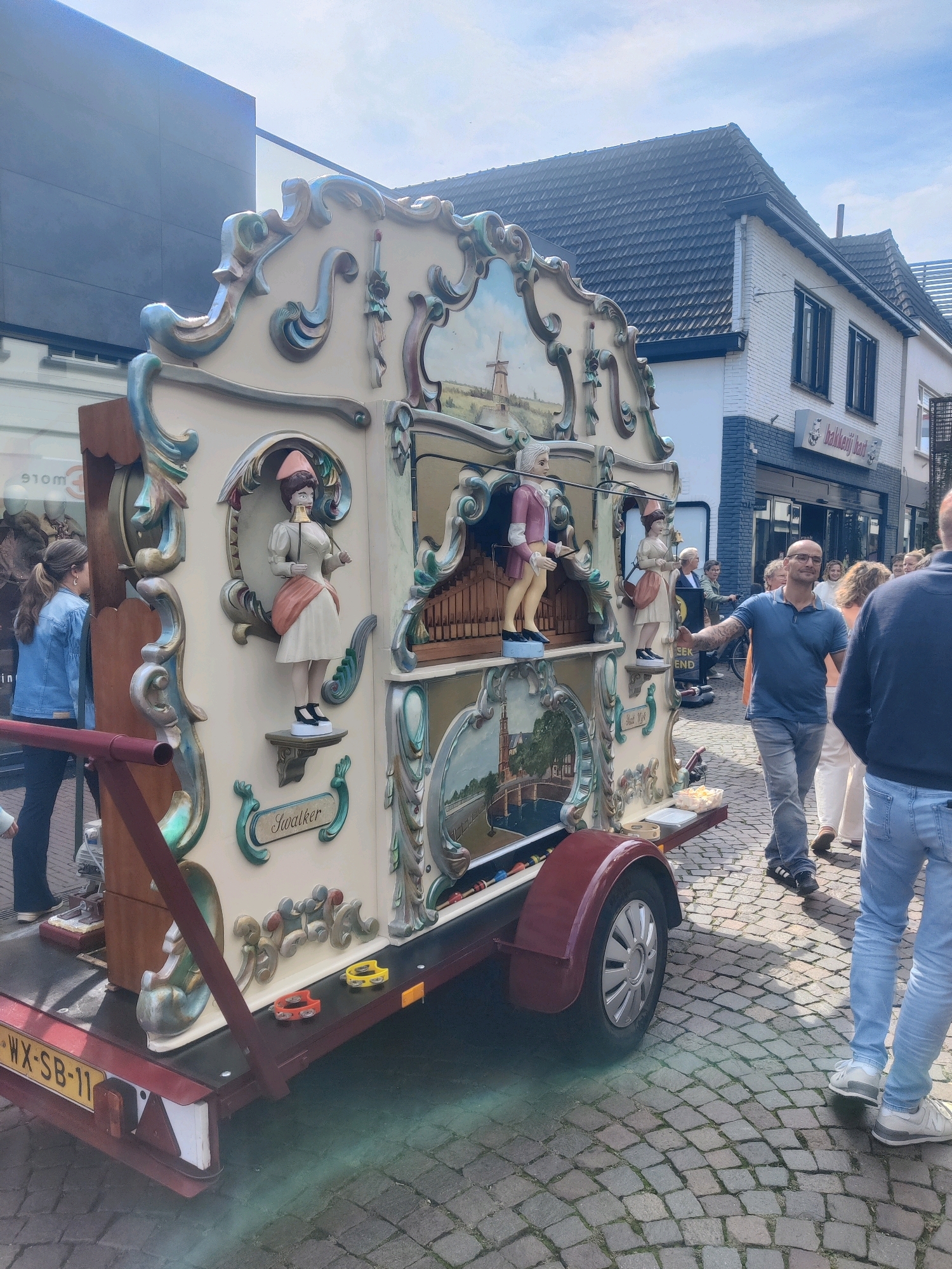 The image captures a street scene in Winterswijk, Netherlands, featuring a large, elaborately decorated barrel organ on a trailer. The barrel organ is cream-colored with ornate golden trim and features painted scenes and three animated figures: a woman in a pink hat and white dress, a man in a pink jacket and white pants, and another woman in a pink hat and white dress, each holding an instrument. The name "Swaalker" is inscribed on a decorative plaque. The barrel organ is parked on a cobblestone street.
In the foreground, the license plate of the trailer reads "WX-SB-11". A man in a blue polo shirt and grey pants stands to the right of the barrel organ, smiling and gesturing outwards with his right arm. To his right, two individuals are partially visible, one in a blue denim jacket and the other in a dark jacket and jeans. In the background, a white building with a tiled roof and black window frames is visible, with a sign that reads "bakkerij hart". Another building to the left has a dark facade and large windows. Several other people are seen in the background, some appearing to be walking away from the barrel organ. The sky is partly cloudy and bright, suggesting daytime.