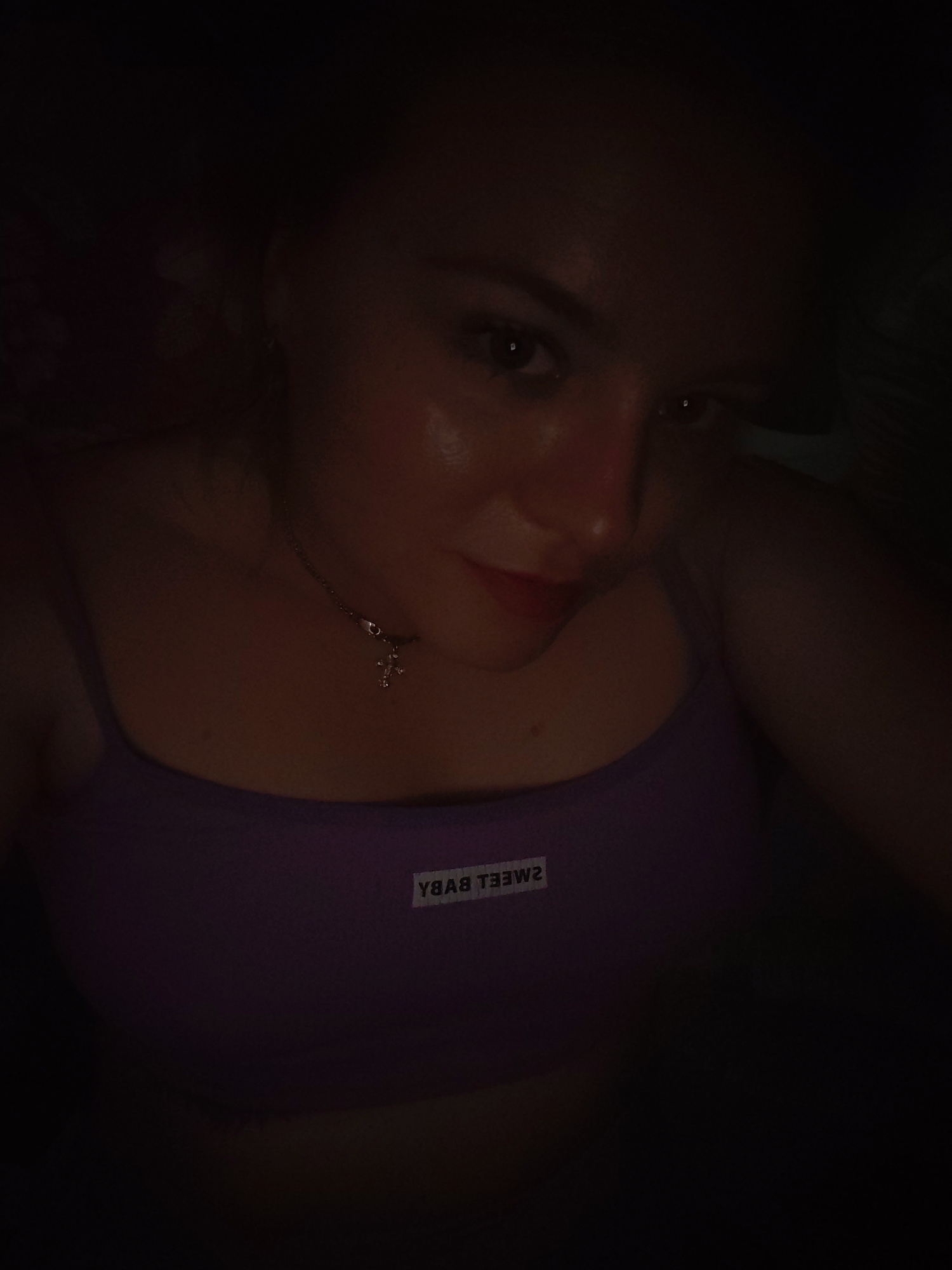 A selfie of a woman wearing a purple tank top with the text "SWEET BABY" reversed on a white label. She is wearing a necklace with a cross pendant. The location cannot be determined from the image.
