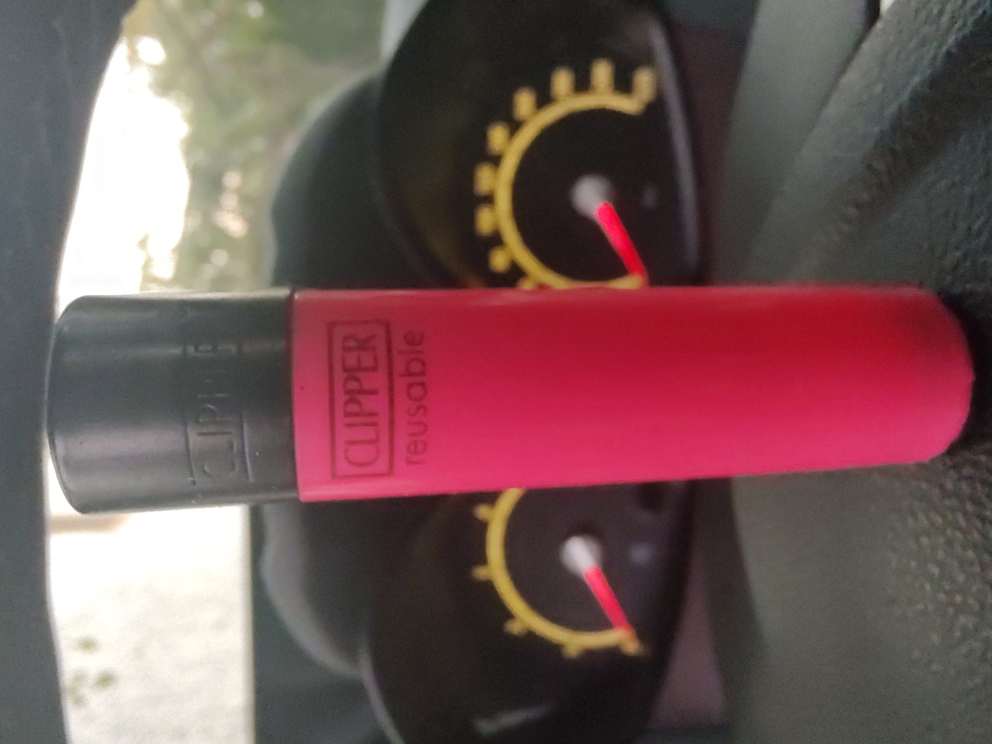 A bright pink Clipper brand lighter with a black cap rests vertically on the dashboard of a vehicle. The lighter displays the word "CLIPPER" in white text within a black rectangle, and below it, "reusable" is also written in white text. The background of the image is out of focus, showing the blurry outlines of a car's dashboard instruments, characterized by yellow semicircular markings and red indicator needles. Beyond the dashboard, the scene transitions to an outdoor environment with blurred greenery and the bright sky suggesting daylight. The image was taken in Tervel, Bulgaria.