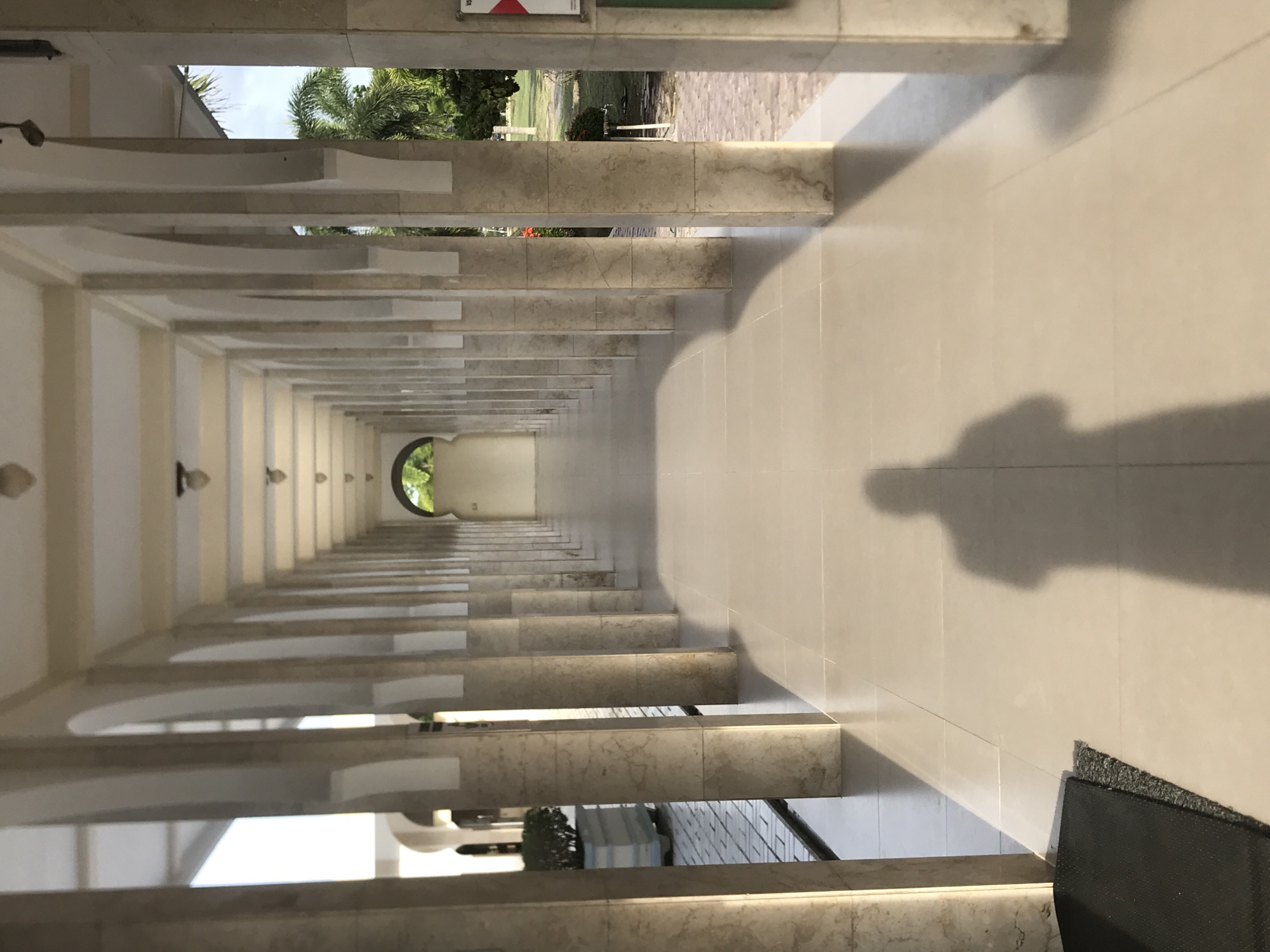 A perspective view of a long, enclosed hallway with evenly spaced archways is shown. Light enters from the end of the hallway and the archways, creating a sense of depth. The walls are a light color, and the floor is tiled, reflecting the light. A shadow of a person is cast on the floor, implying someone is present, but they are not visible. Outside the archways, hints of green foliage and what appears to be a lawn can be seen. The location is Tambontunong, Indonesia, as stated.