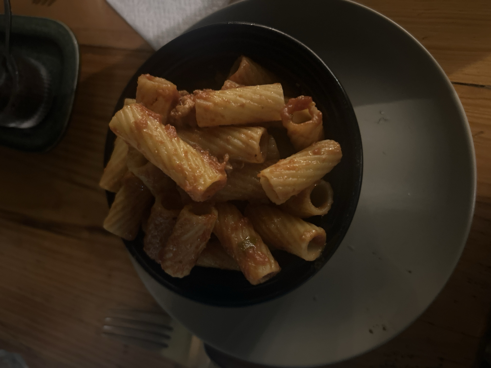 A close-up shot depicts a bowl of rigatoni pasta with tomato sauce, presented on a gray plate. The bowl is black. A portion of a glass and a napkin are visible in the upper-left corner. The pasta dish is set on a wooden table. A fork lies near the edge of the plate. The location is potentially San Bernardino Tlaxcalancingo, Mexico.