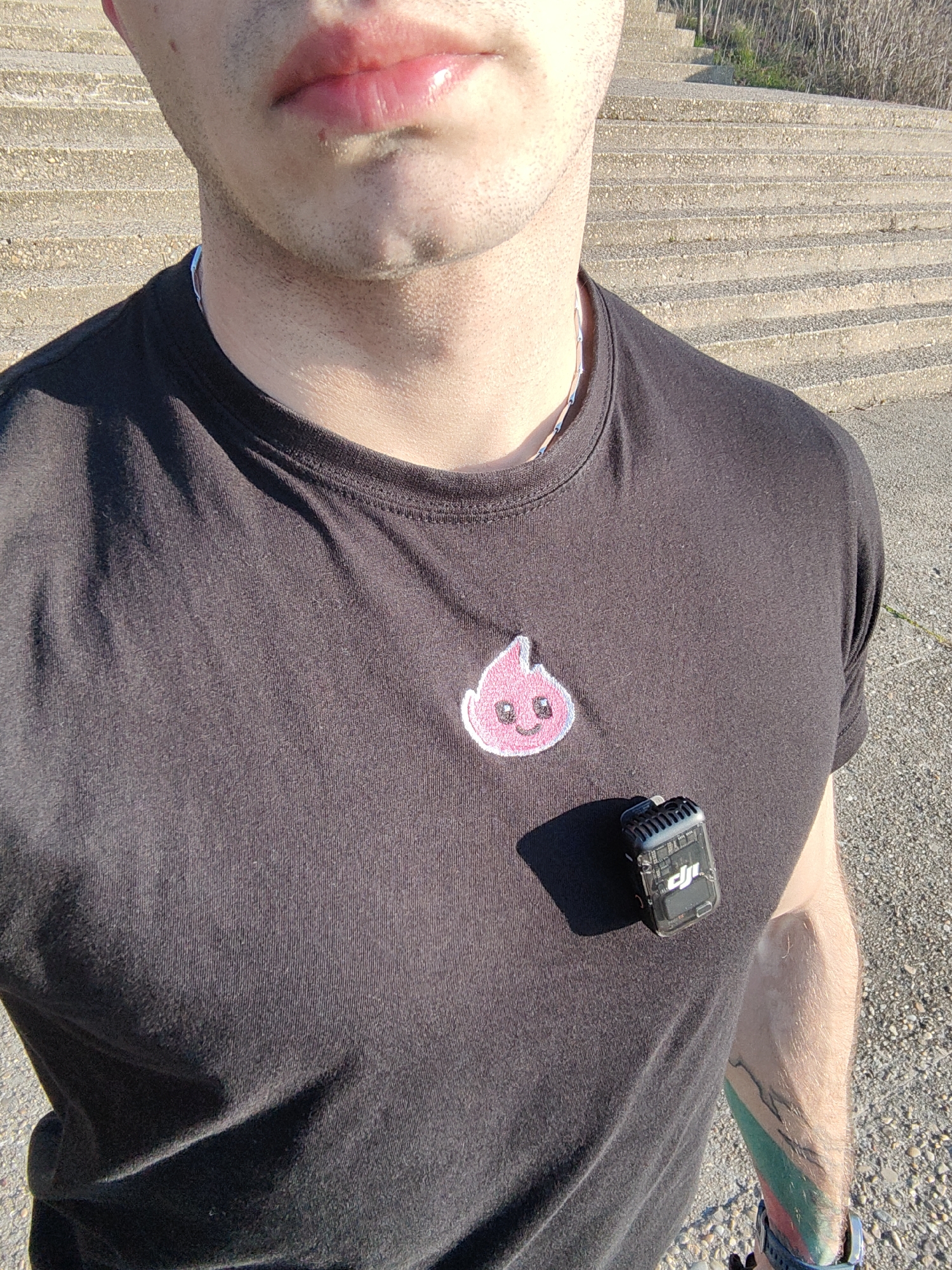A male individual is partially visible, facing forward, from the upper chest to just below the chin, with part of his right arm also shown. He has short facial stubble and full, hydrated lips. He wears a dark, likely black, short-sleeved t-shirt with a thin silver-colored chain around his neck. On the left chest area of the t-shirt, an embroidered patch depicting a pink, smiling, drop-shaped character is present. Clipped to the right chest area is a black and transparent DJI wireless microphone transmitter, casting a distinct shadow. The individual's right forearm features a multi-colored tattoo, including green and red hues, and a blue wristband or smartwatch is visible on his right wrist. The scene is outdoors, illuminated by direct sunlight, creating sharp shadows. In the background, a wide flight of light grey concrete steps, textured with aggregate, ascends diagonally from left to right. Sparse, dry vegetation is visible alongside the upper section of the steps. The ground surface in the foreground appears to be light-colored gravel or asphalt. The location is an outdoor area with concrete steps, possibly within a public park or elevated section of the city, such as the Sea Garden in Varna, Bulgaria.